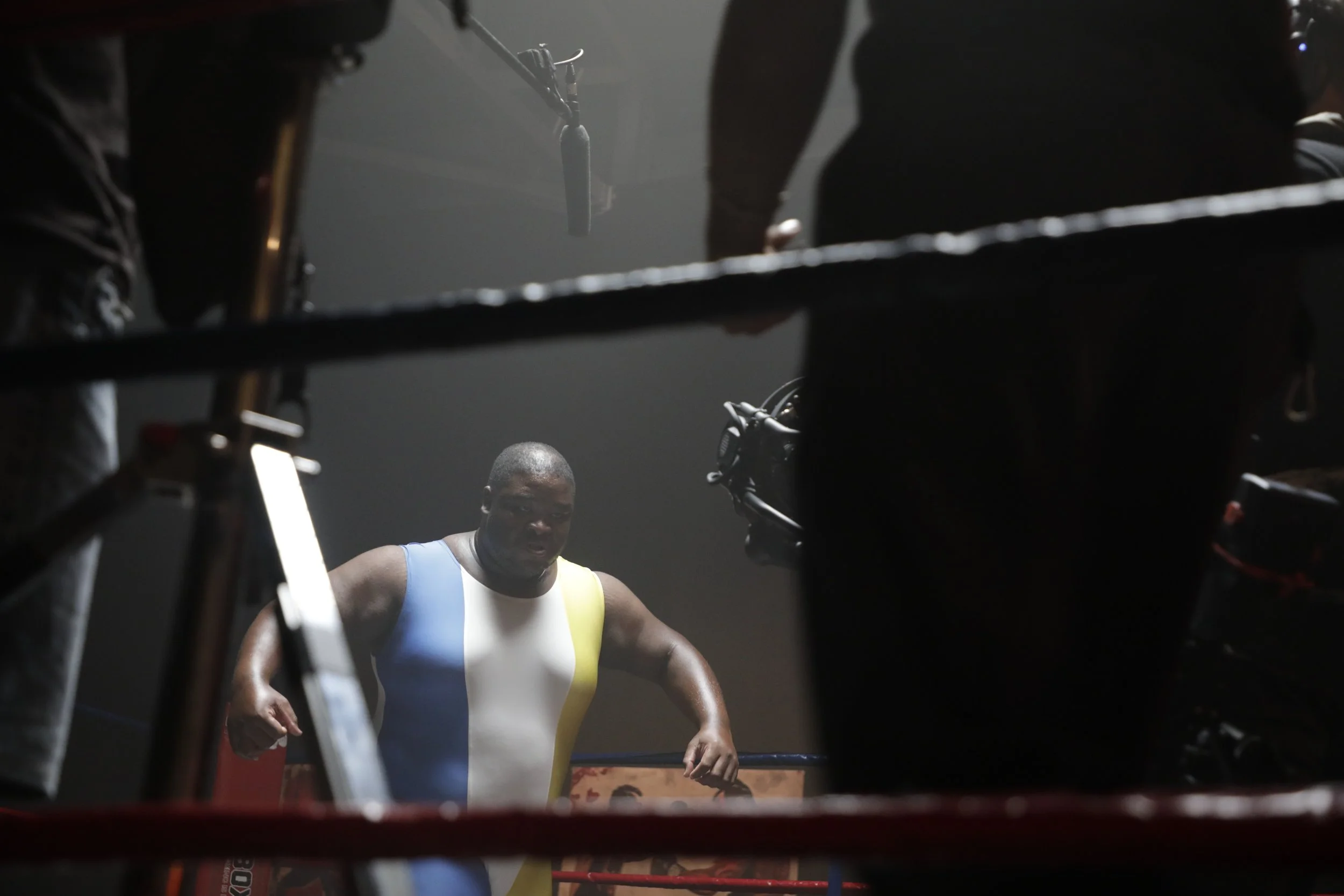 A boxer in a sleeveless uniform with blue, white, and yellow colors standing inside a boxing ring, surrounded by people, with a dark background.
