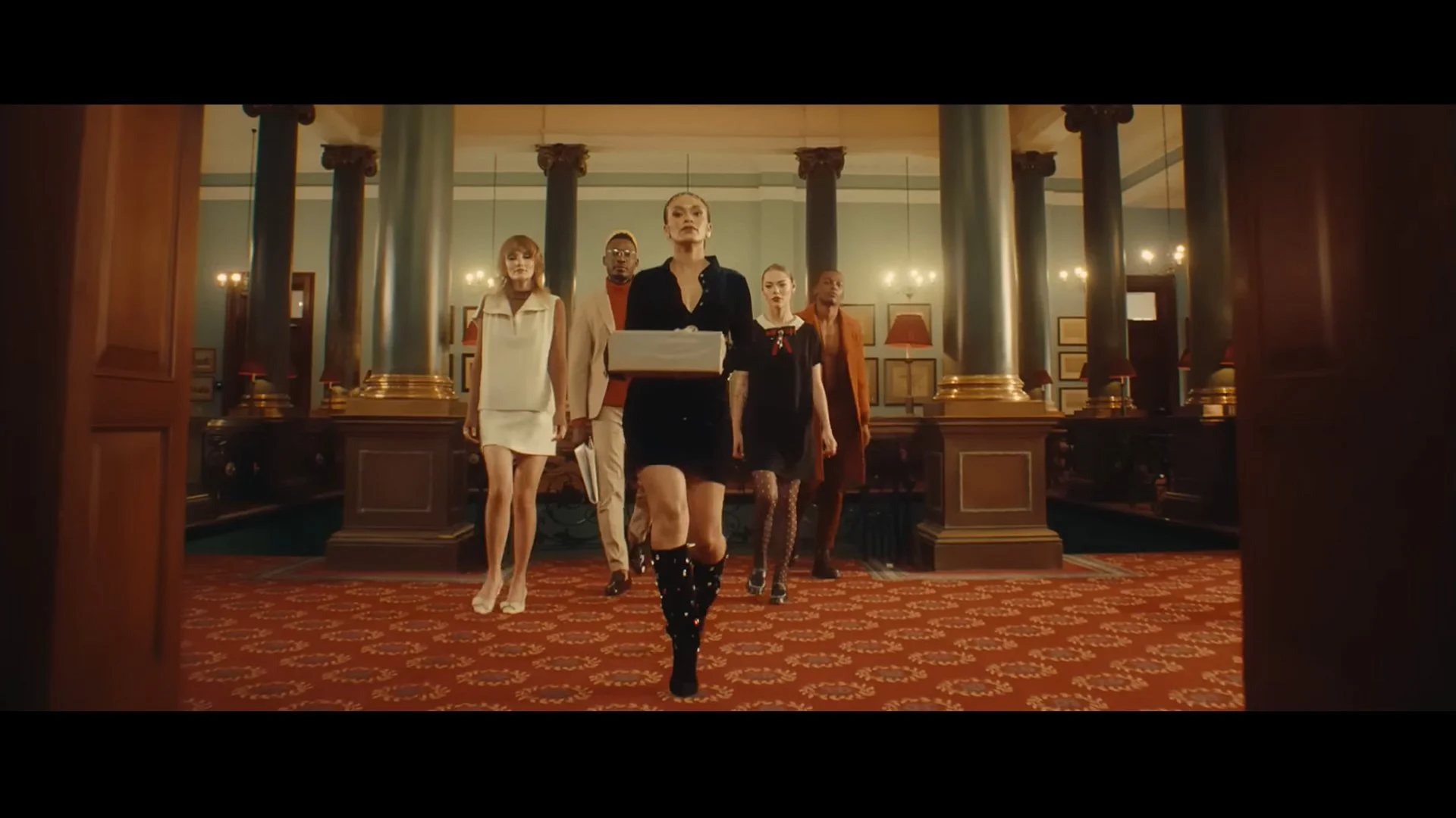 A group of five fashionable people walking through a grand, ornately decorated room with tall dark columns, warm lighting, and framed artwork on the walls.