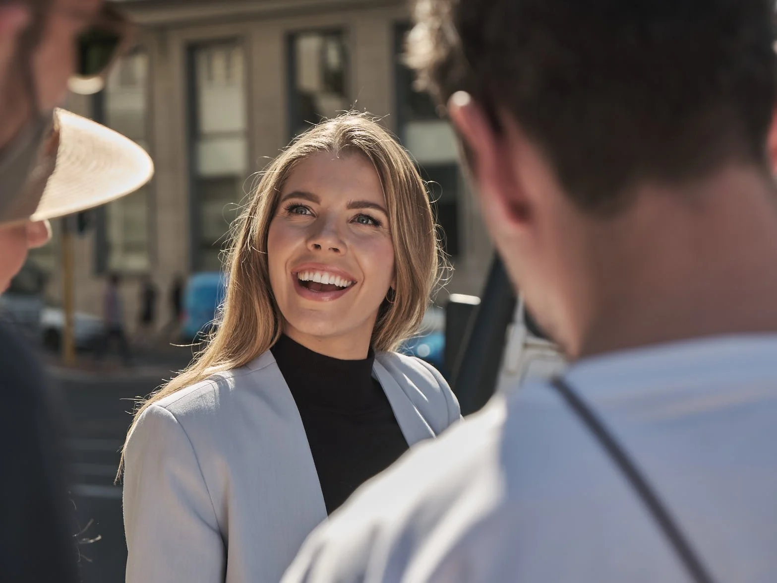 A smiling woman engaged in conversation outdoors with two men, one wearing a hat and the other with dark hair, in a city setting.