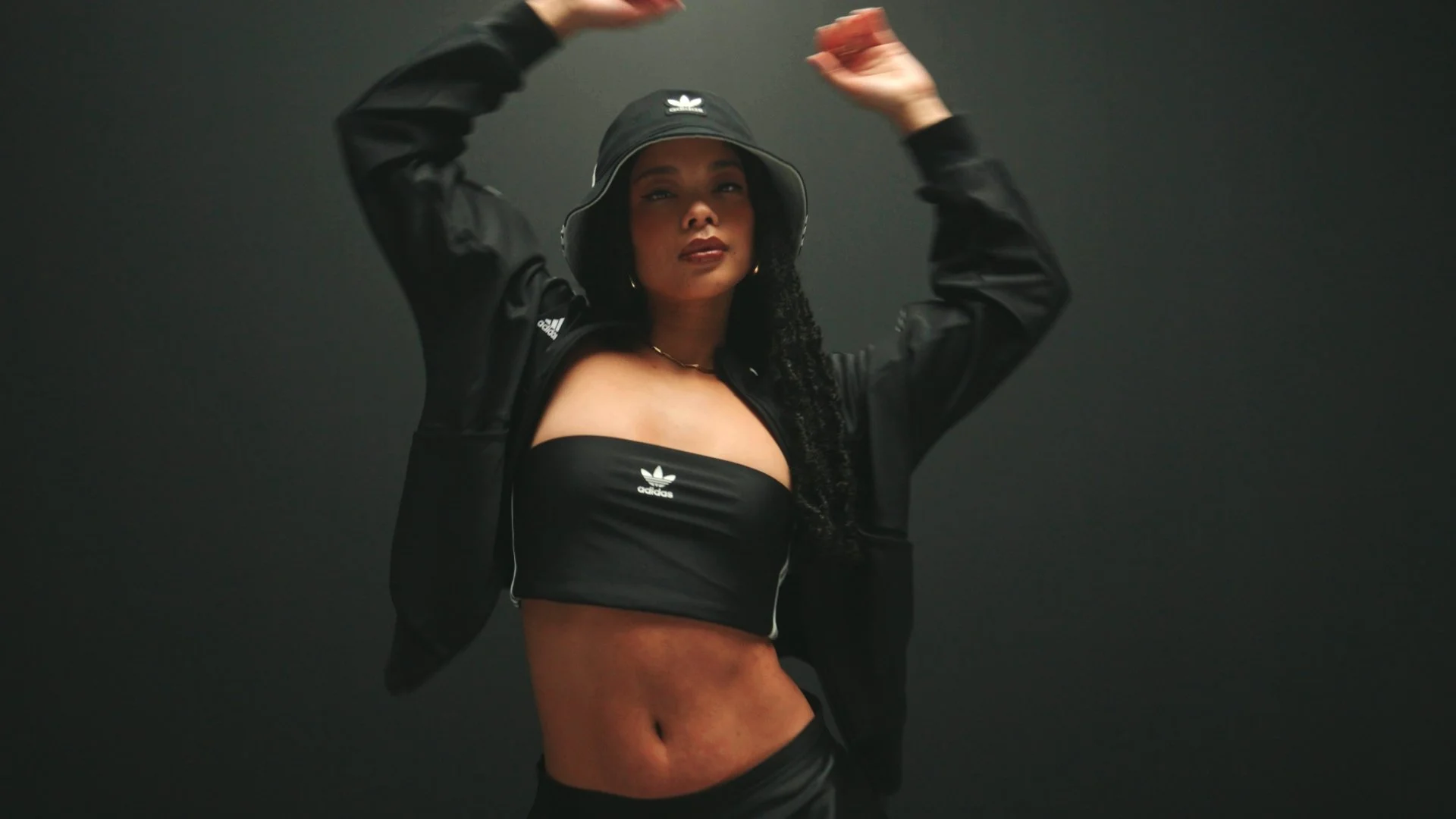 Woman wearing black Adidas sportswear and bucket hat, with her arms raised, posing against a dark background.