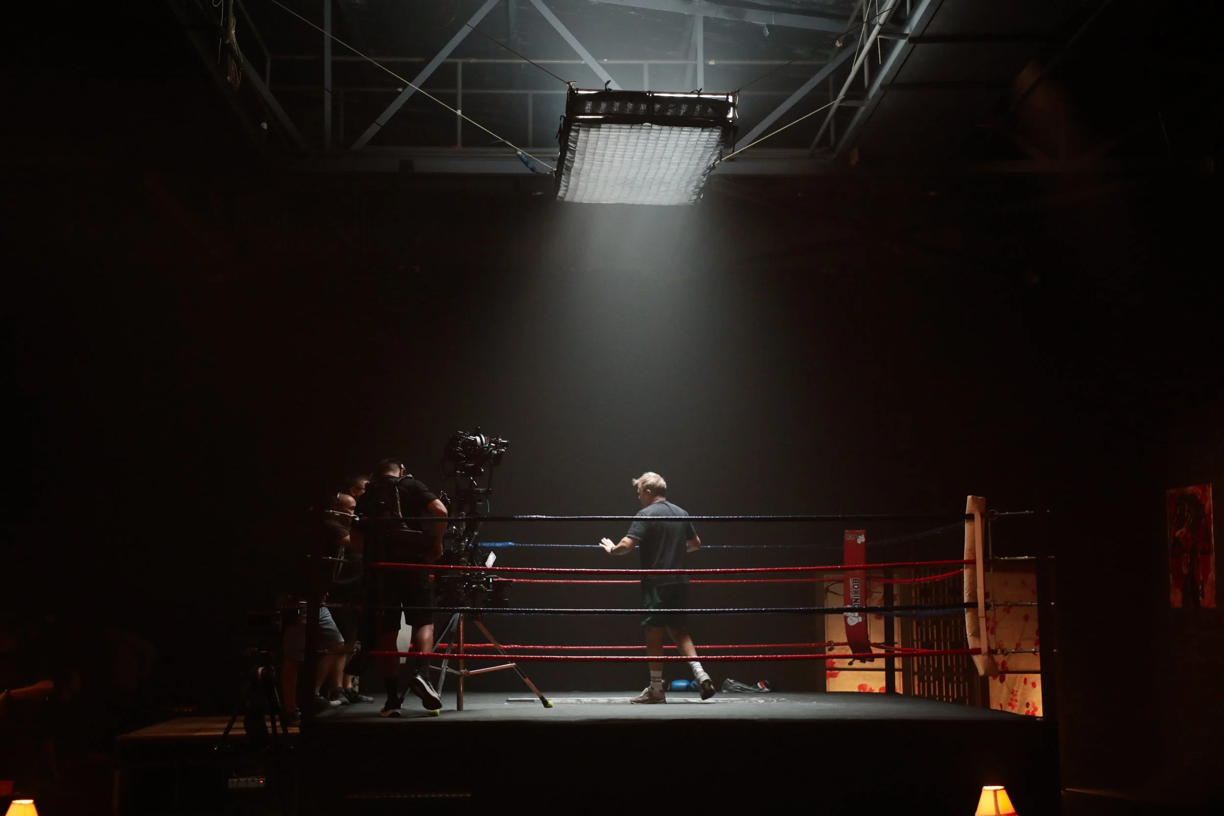A boxing ring inside a dark gym with four men, one in the ring, and three around it, one filming and two talking.