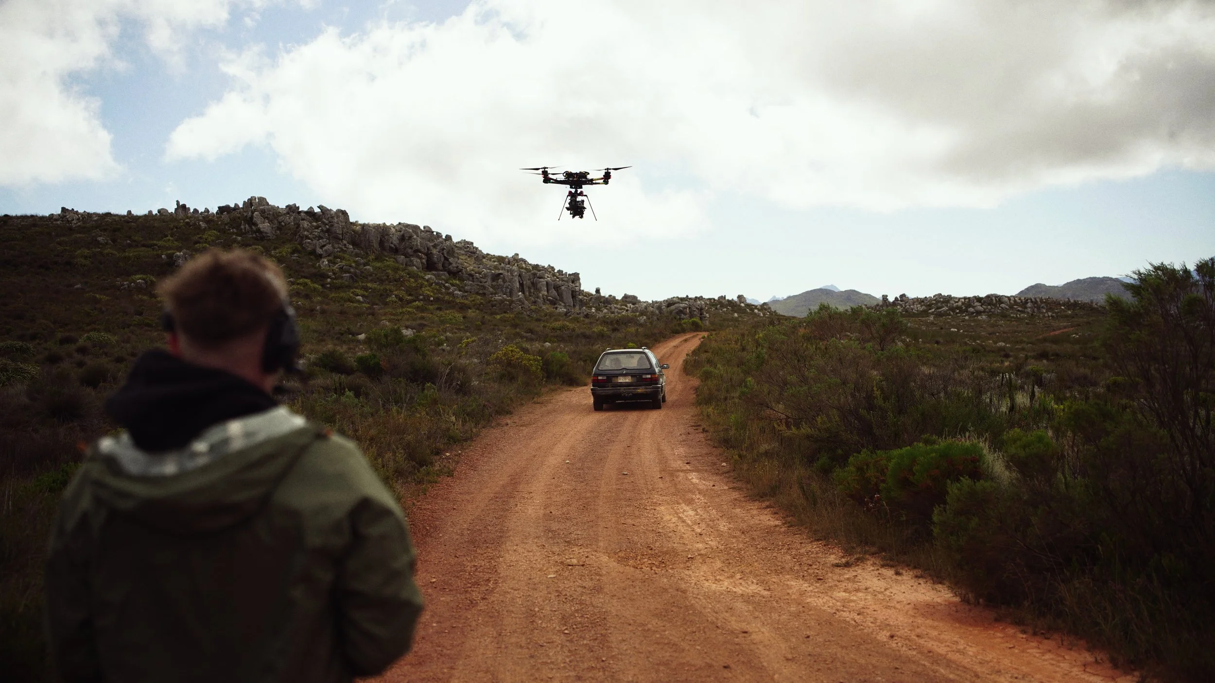 A person wearing a jacket and headphones stands on a dirt road in a rural area, observing a drone flying overhead with a camera attached, with a car driving ahead on the road and mountains and clouds in the background.