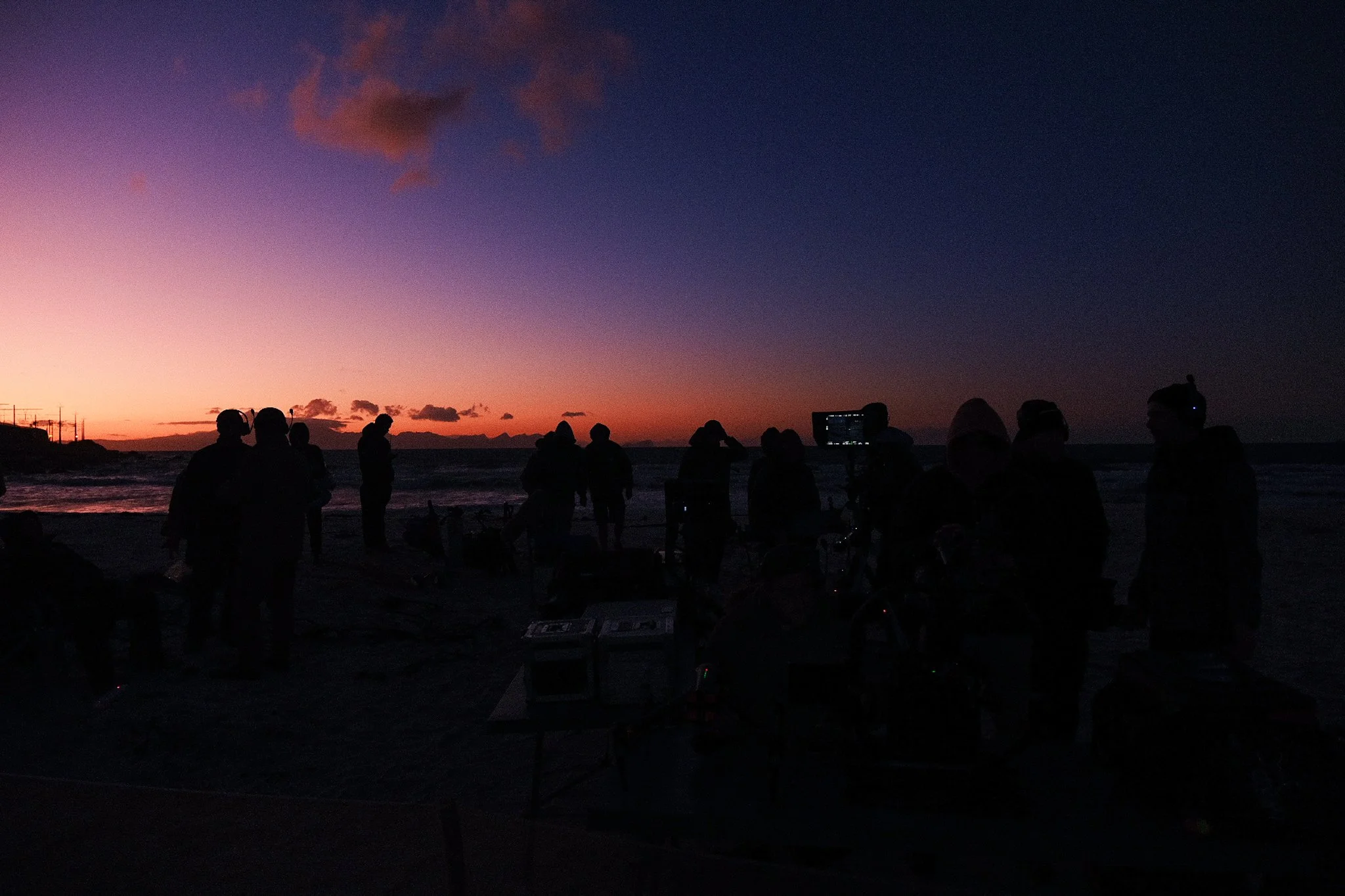 Silhouetted people on a beach during sunset, some with equipment, with a colorful sky transitioning from pink to dark blue.