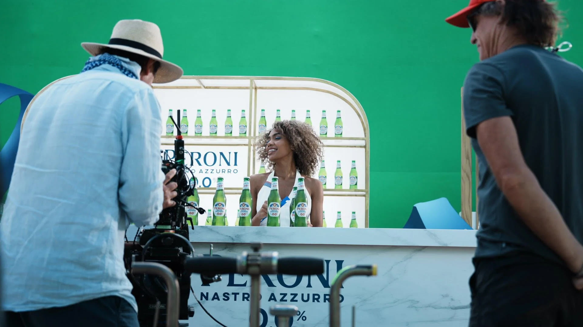 A woman with curly hair smiles behind a bar with bottles of Peroni Nastro Azzurro beer, while two men film her in front of a green screen backdrop.