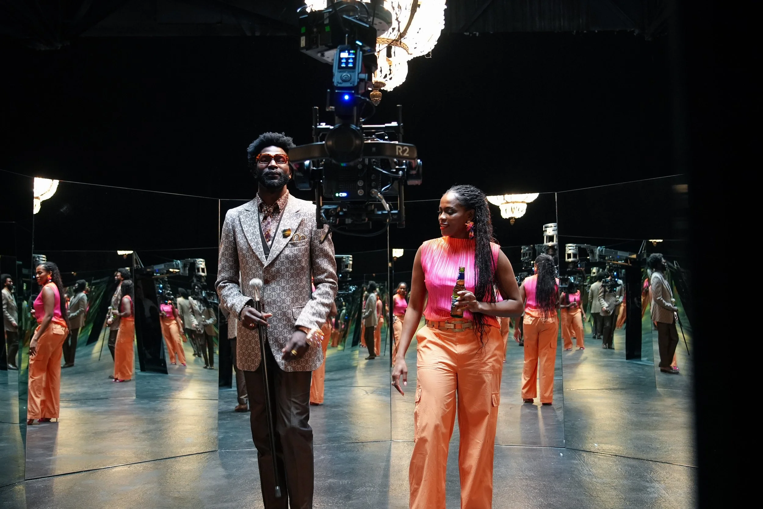 Two people in a room with mirrors, one man wearing a patterned suit and holding a cane, and one woman wearing a pink sleeveless top and orange pants, holding a bottle, surrounded by multiple camera operators and mirrors reflecting their images.