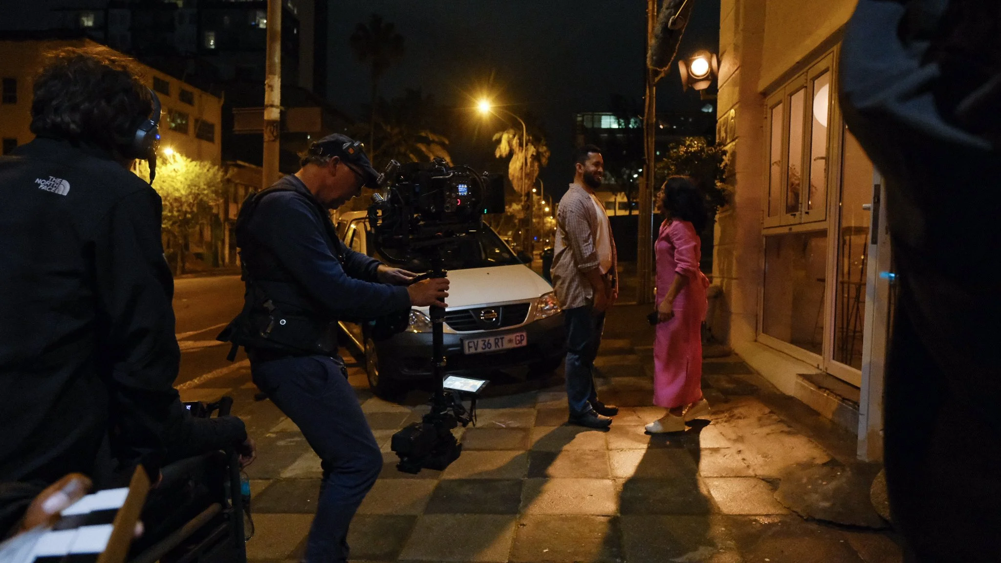 Film crew filming scene on a city street at night with two actors standing outside a building, one actor in a gray shirt and the other in a pink dress, with a camera operator capturing the scene.
