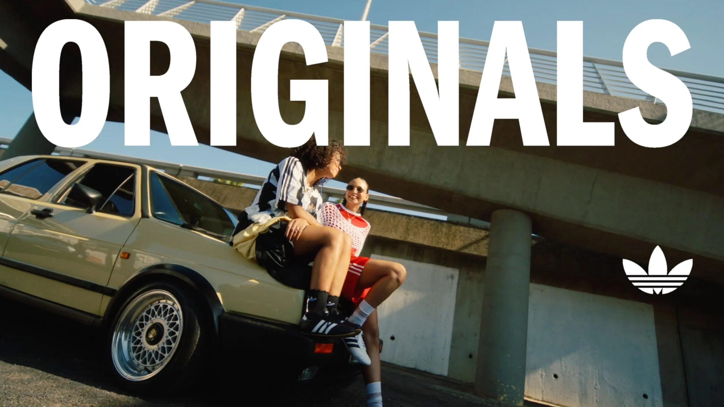 Two women sitting on the hood of a beige vintage car under a bridge, smiling and talking. Large white text reading 'ORIGINALS' overlays the image, with a small Adidas logo in the bottom right corner.