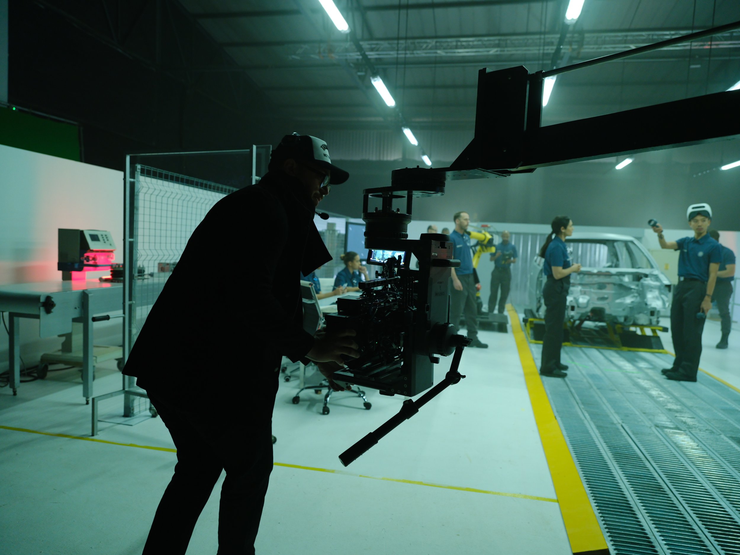 Filmmaker filming a group of engineers working on a car prototype in a laboratory or industrial setting.