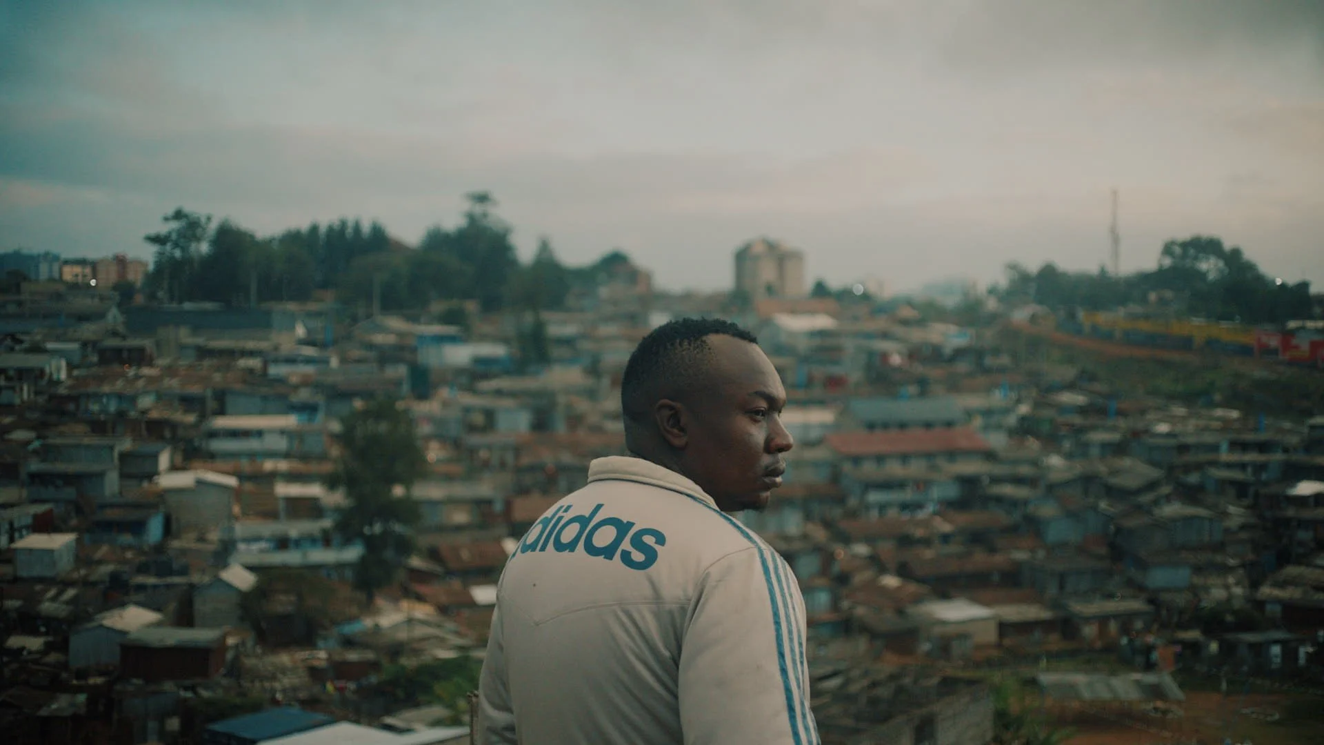 A man in a white Adidas jacket looking over a densely populated urban area with small houses and buildings under a cloudy sky.