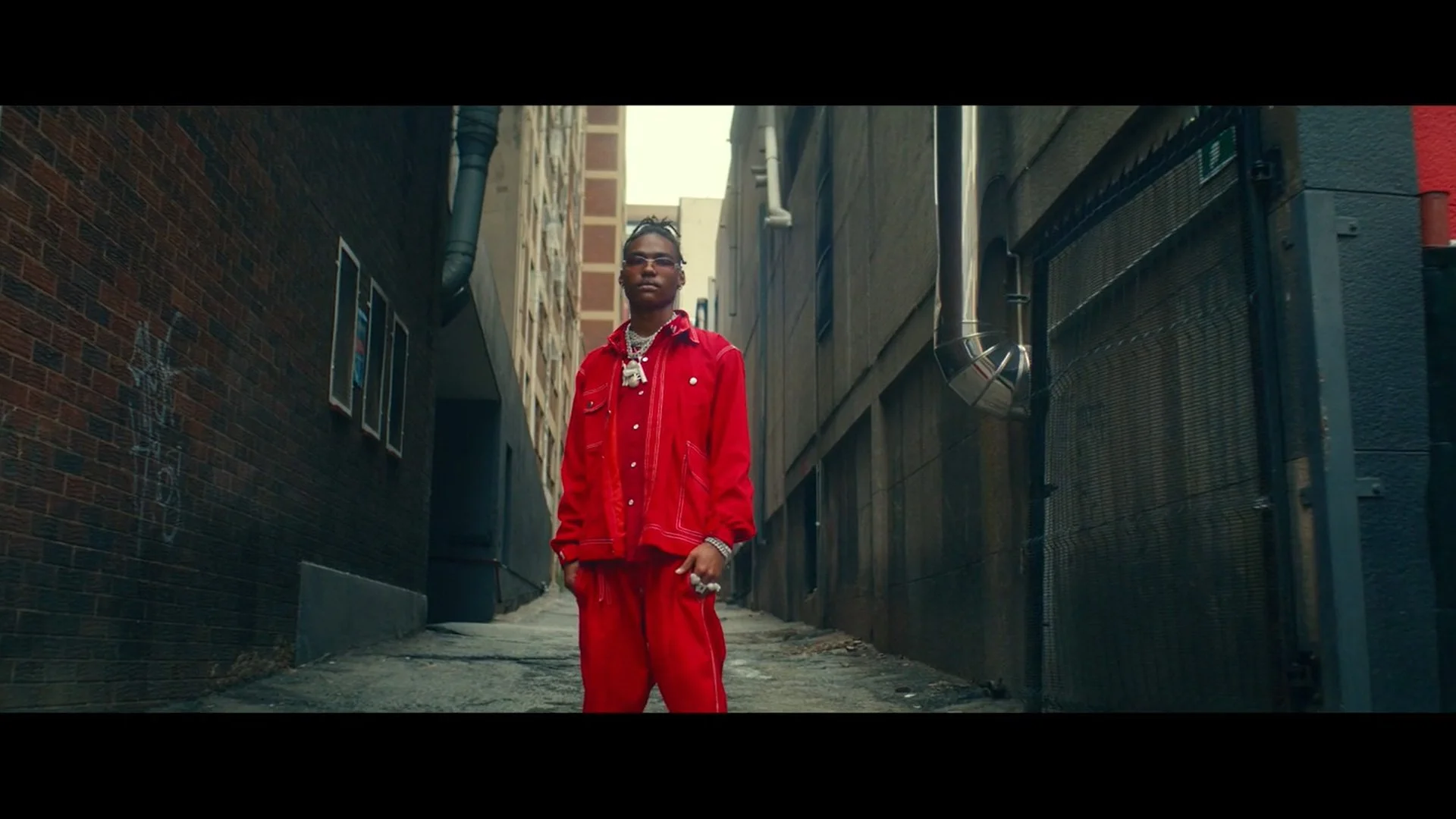 A young person standing in a narrow urban alleyway wearing a red jacket and matching red pants, with jewelry and sunglasses, looking confidently at the camera.