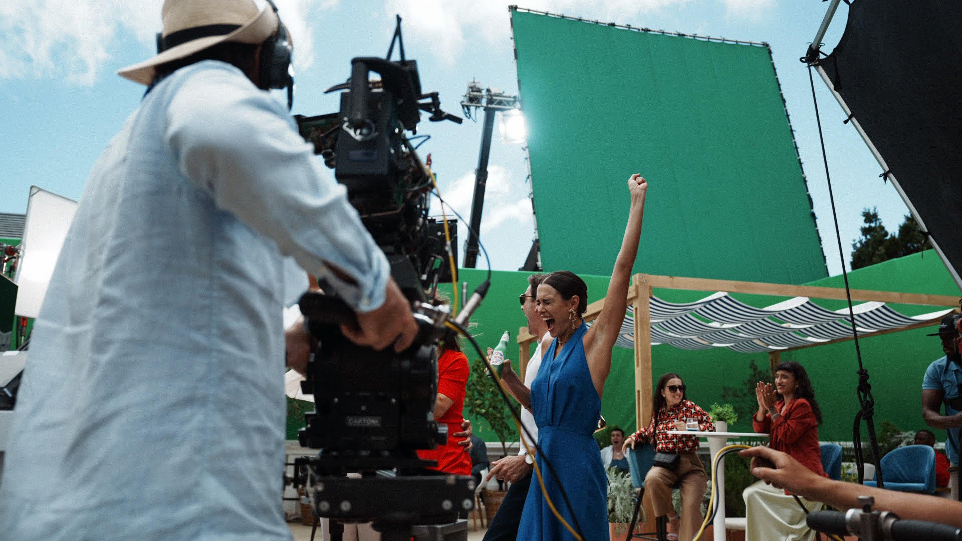 Filming of a lively outdoor scene with a woman in a blue dress celebrating, surrounded by crew and audience, on a set with green screens.