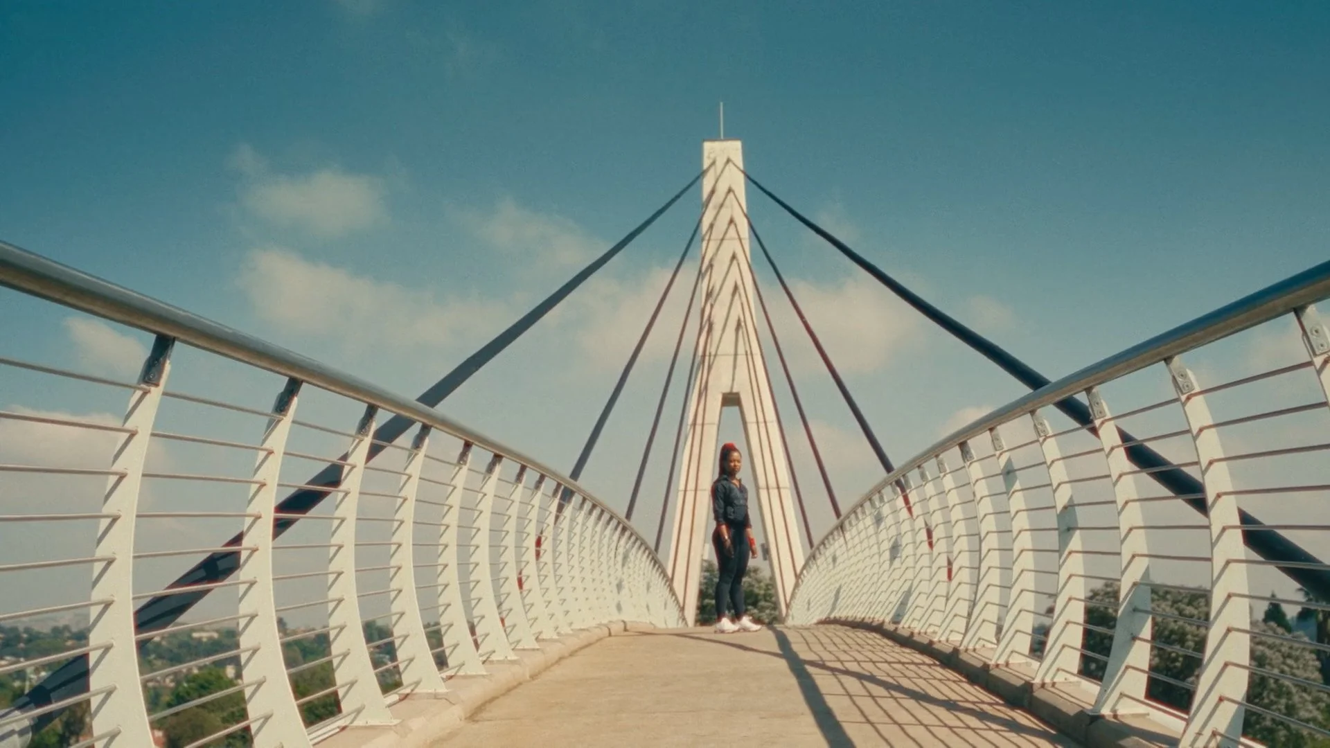 A woman stands on a modern, white suspension bridge with curved railings and tall support towers, under a partly cloudy sky.
