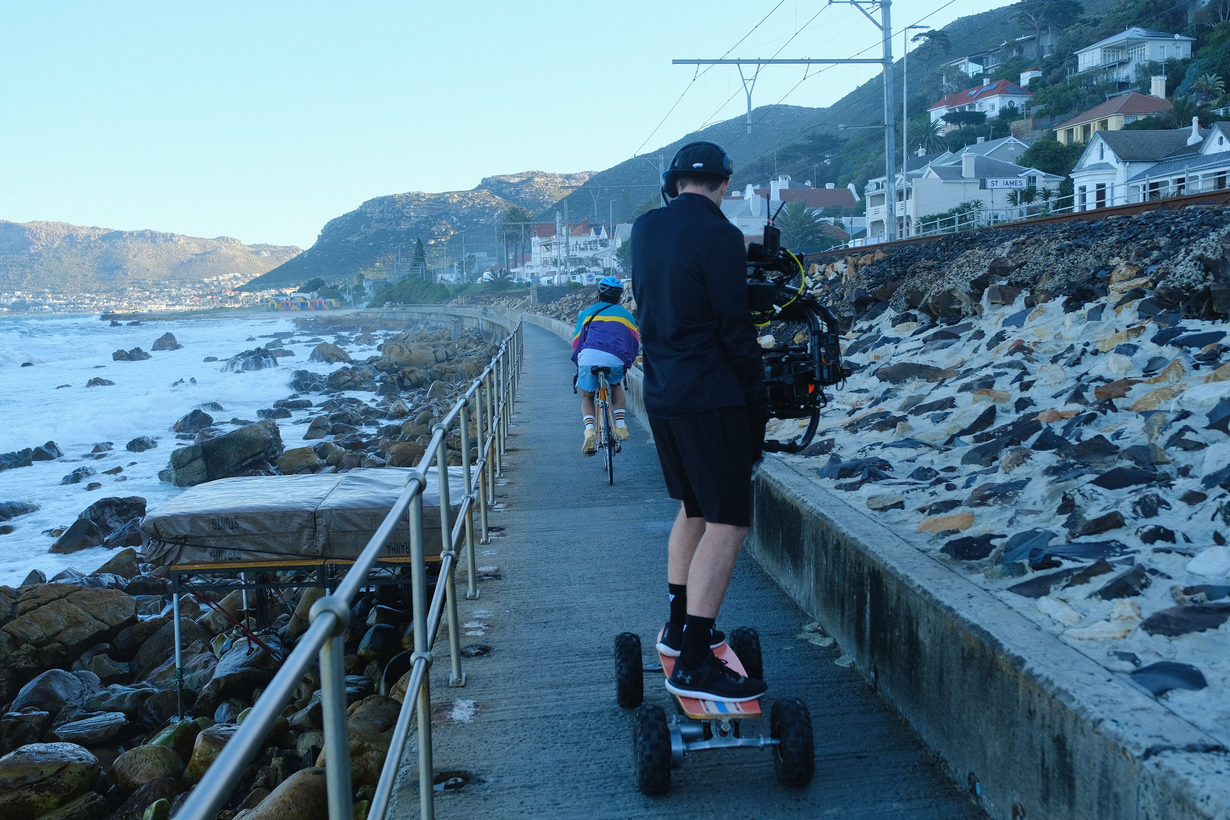 Person filming with a camera on a hoverboard walking along a seaside walkway, with a cyclist riding ahead, rocky shoreline on the left and residential houses on a hillside in the background.