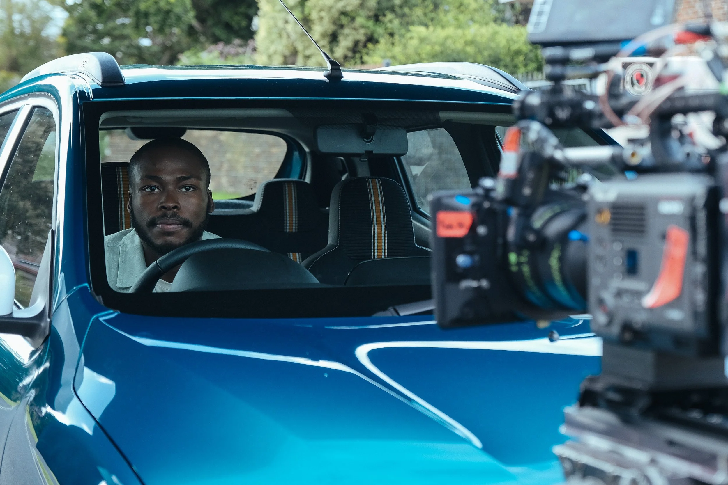 Man sitting inside a blue car on a film set, looking at a professional camera.