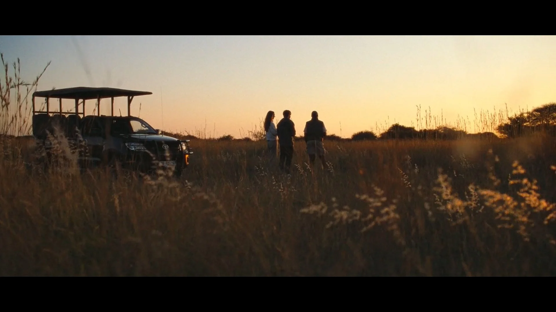 Three people standing and talking in a grassy field during sunset, with a black safari vehicle parked nearby.