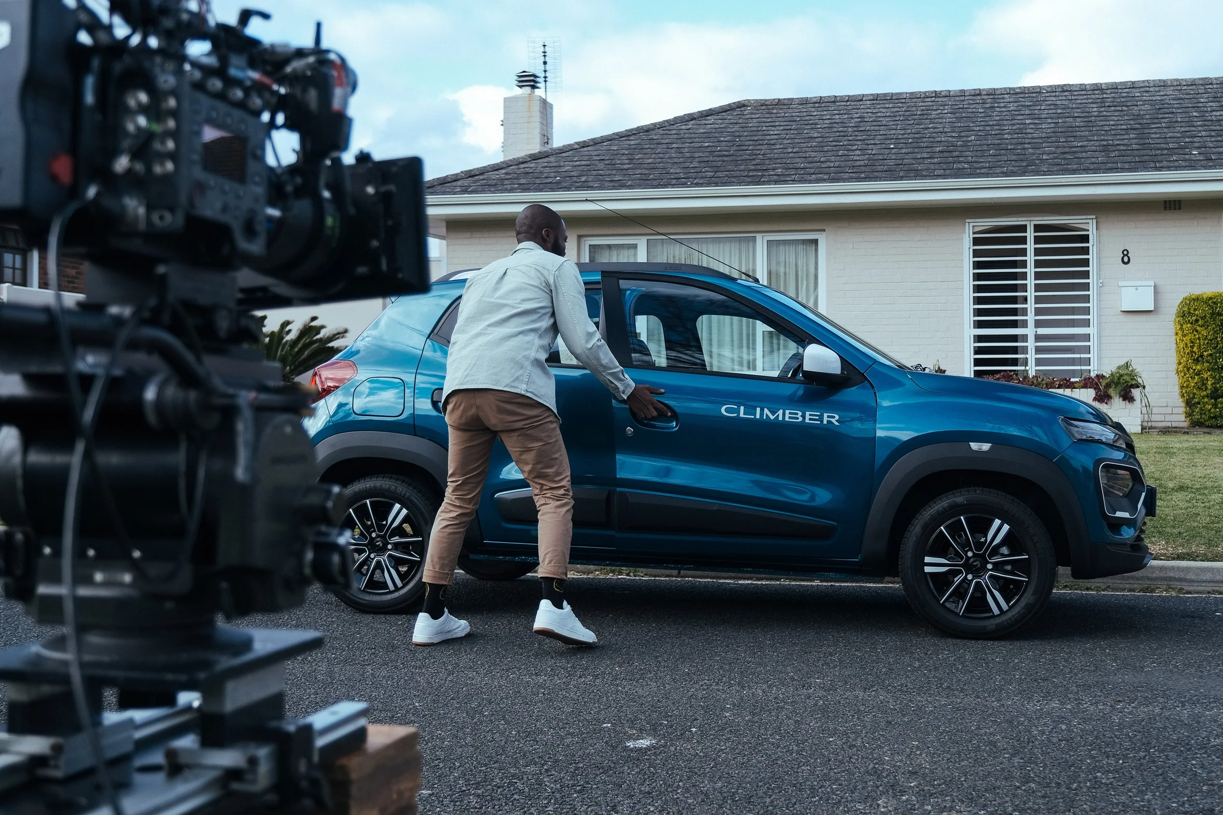 A man is filming a blue Compact SUV with the word 'CLIMBER' on it in front of a house, with camera equipment visible in the foreground.