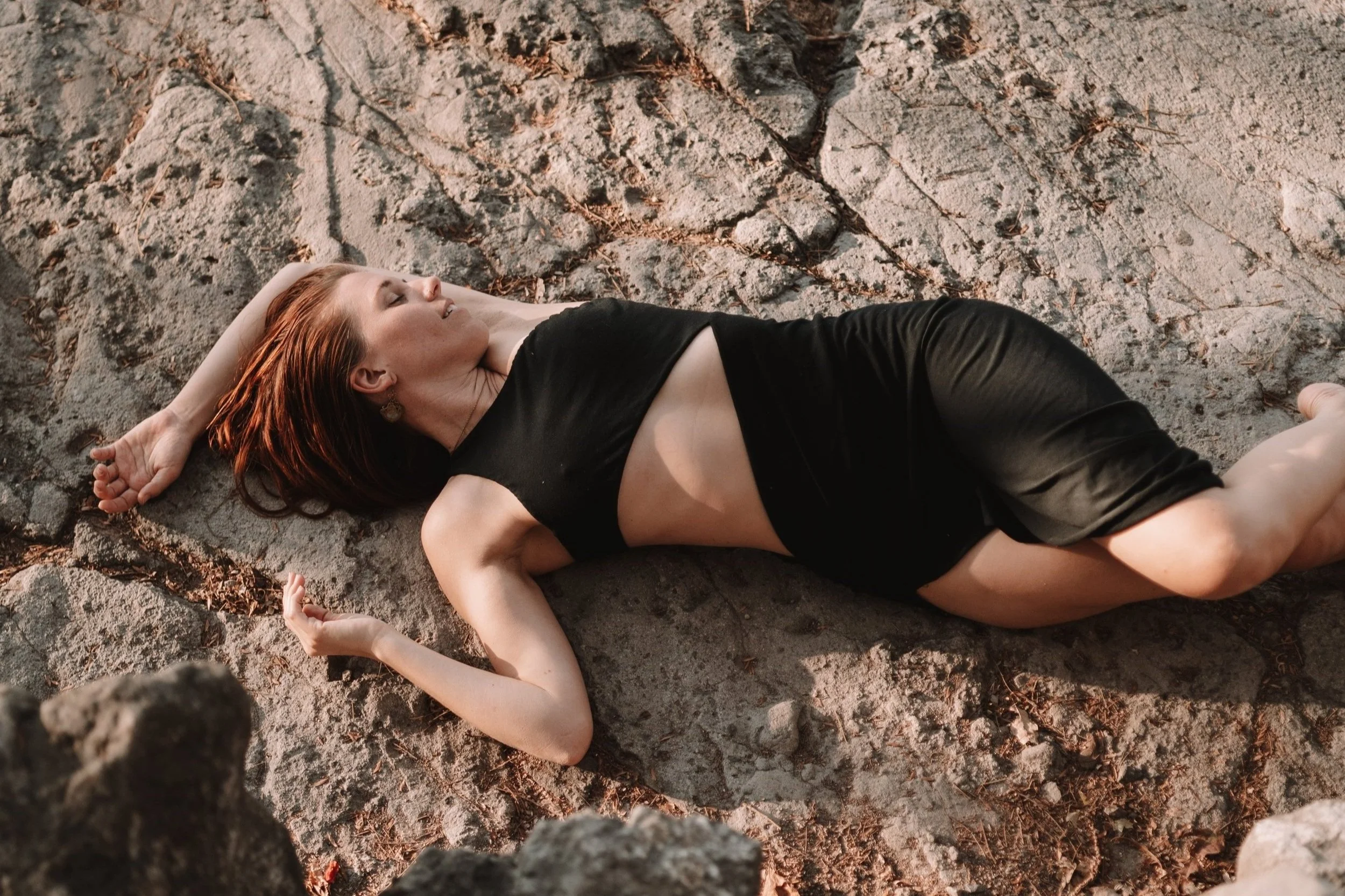 A woman with red hair is lying on a rocky surface with her eyes closed, wearing a black sleeveless dress with a cutout showing her midriff.