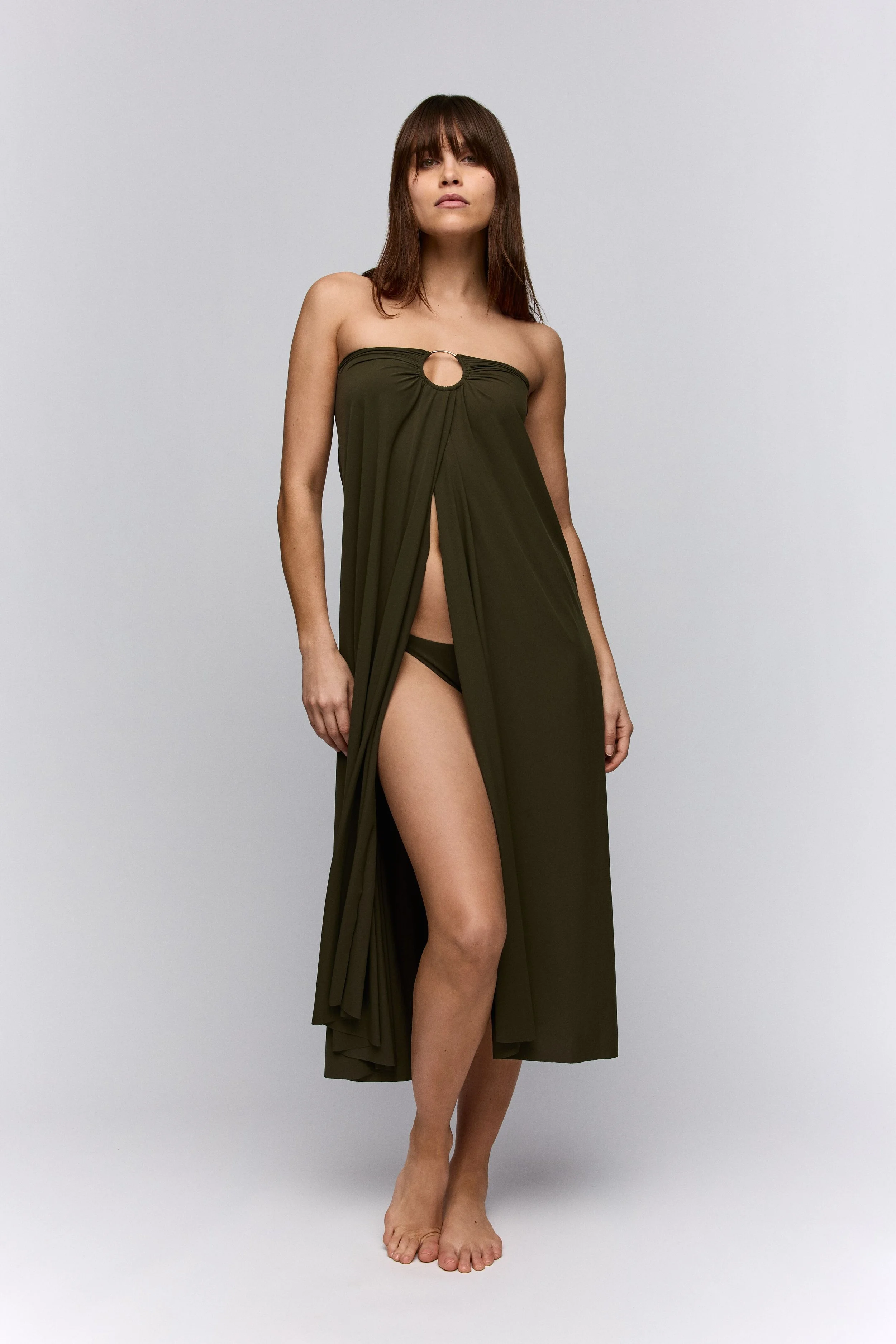 Nainen seisoo paljain jaloin vaalealla taustalla, hänellä on yllään oliivinvihreä ranta-asu ja samanväriset bikinit.