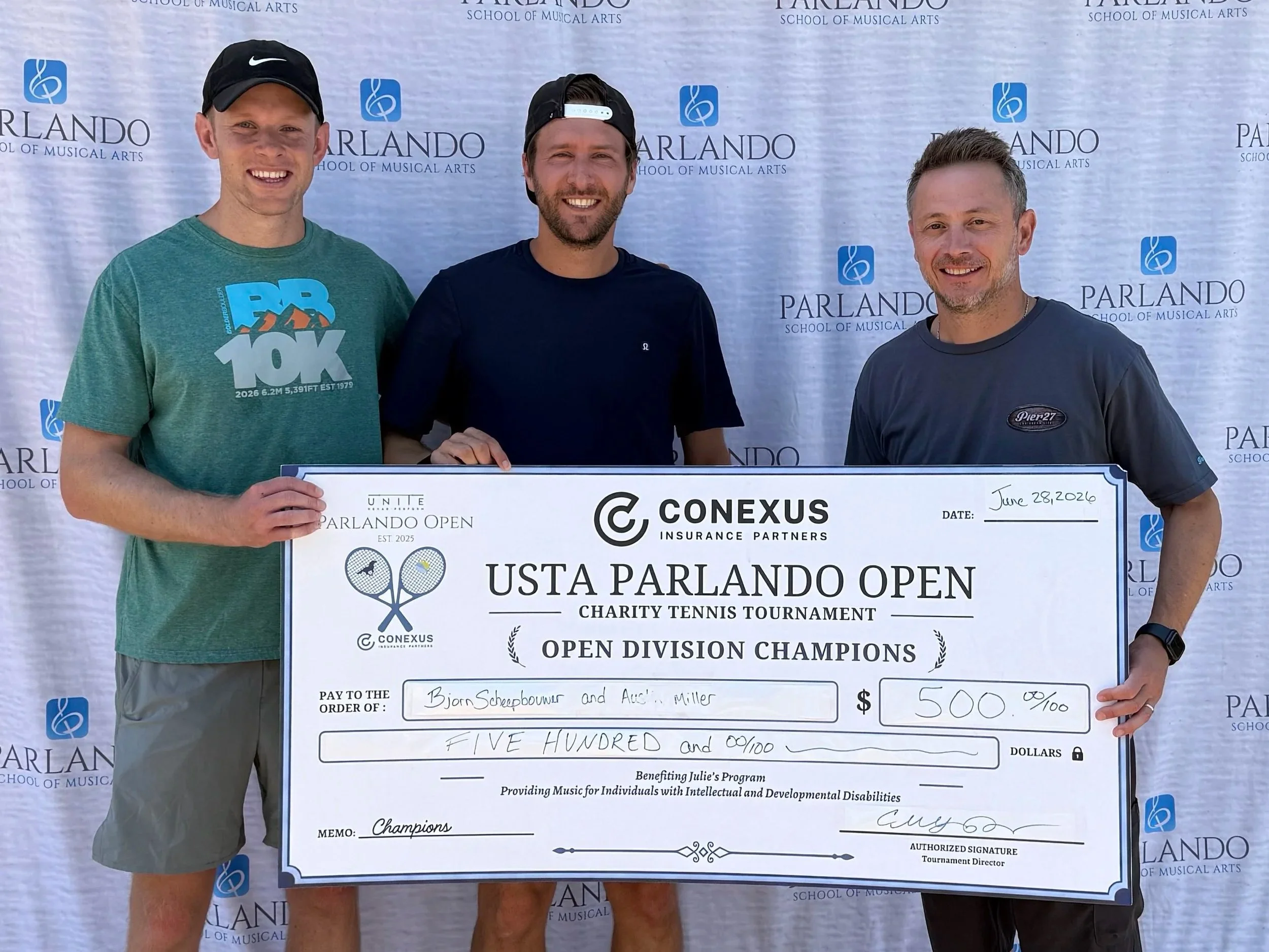 Three men smiling and holding a large check in front of a backdrop with 'Parlando School of Musical Arts' and musical note logos. The check is made out to Bjorn Scheppouwer and Ace Miller for $500, dated June 28, 2026, awarded as champs of a charity tennis tournament.