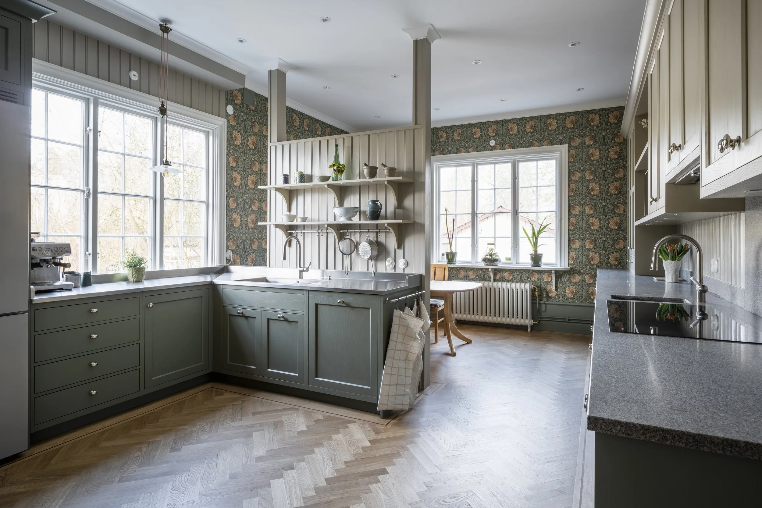 Kjøkken med grønne skap, store vinduer, trebord, blomster, oppheng for kjøkkenutstyr, og blomster på vinduskarmen.