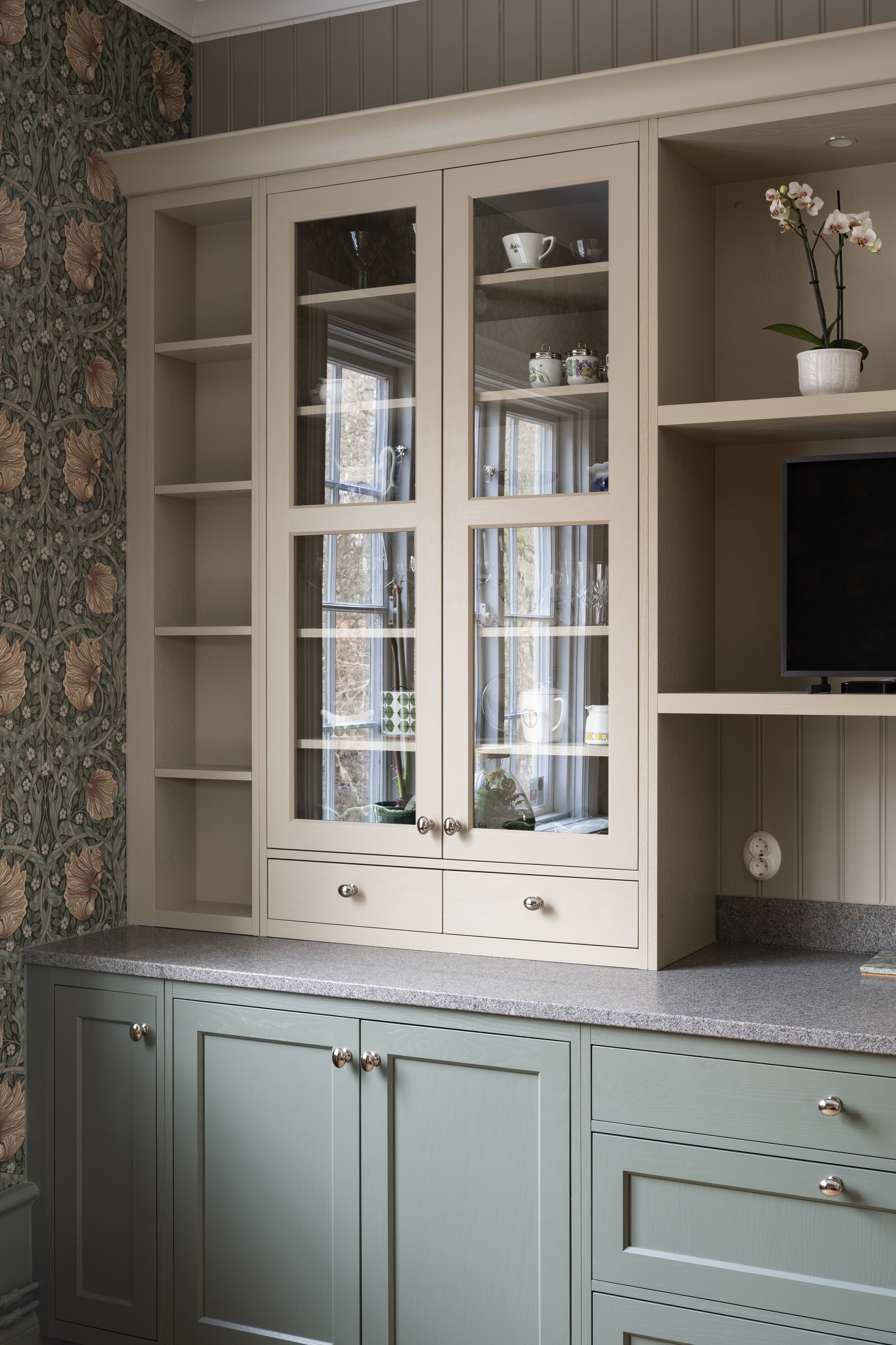 Kjøkkenhyller med glassdører, med keramikk og glassgjenstander. Vegg med floral tapet.