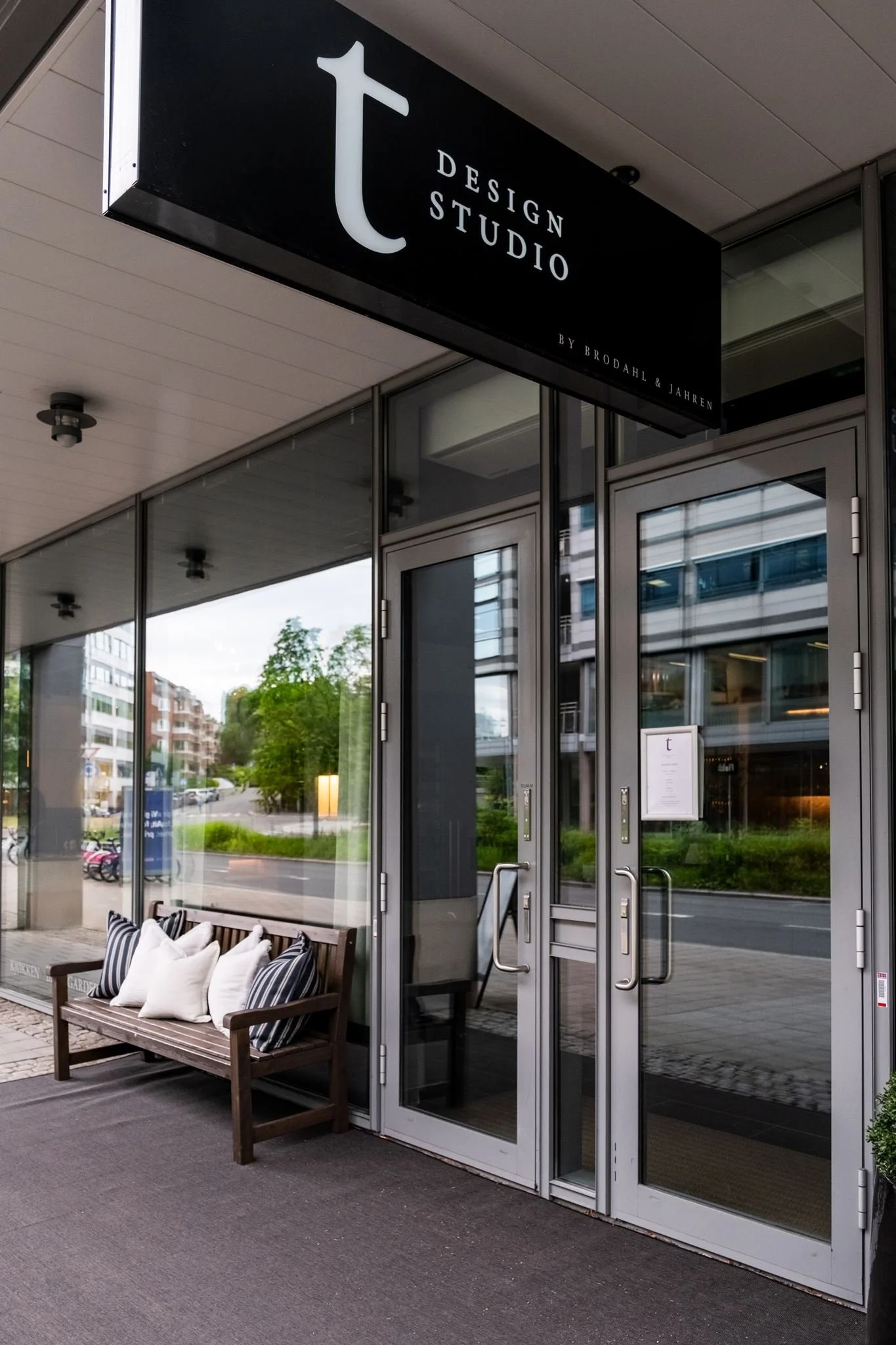 Ute foran en glassdør med en svart skilt som sier 'Design Studio' og en benk med puter utenfor.