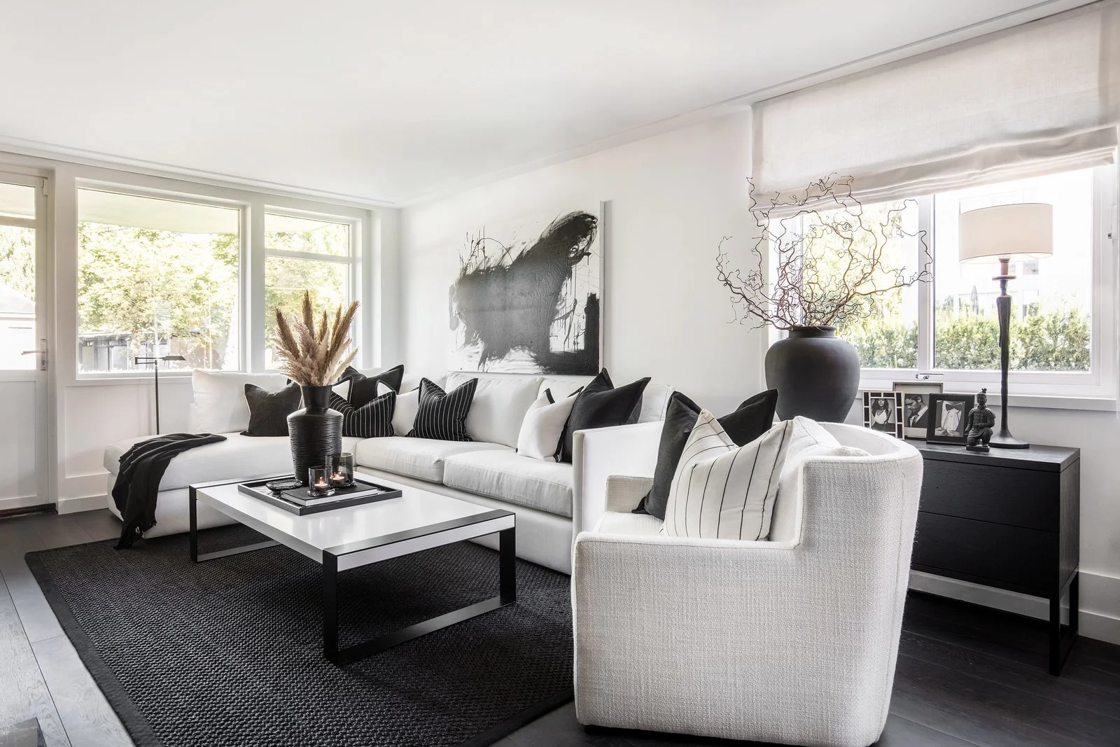 En moderne stue med hvit sofa, svart og hvit puter, svart og hvitt teppe, svart salongbord, stor svart vase med tørre grener, svart sidebord med rammer og dekorasjoner, store vinduer med hvite rullegardiner, svart kunstverk på veggen, svart gulvlampe og bordlampe.