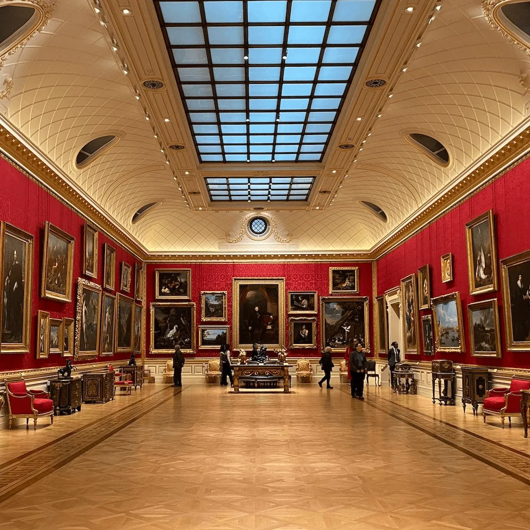 Elegant museum gallery (Wallace Collection) with high ceiling, red walls, and numerous framed paintings. Visitors are observing the art displayed on walls and in center. TRANS VOICES delivered a bespoke set as part of Sadie Lee's LGBTQ+ Late.