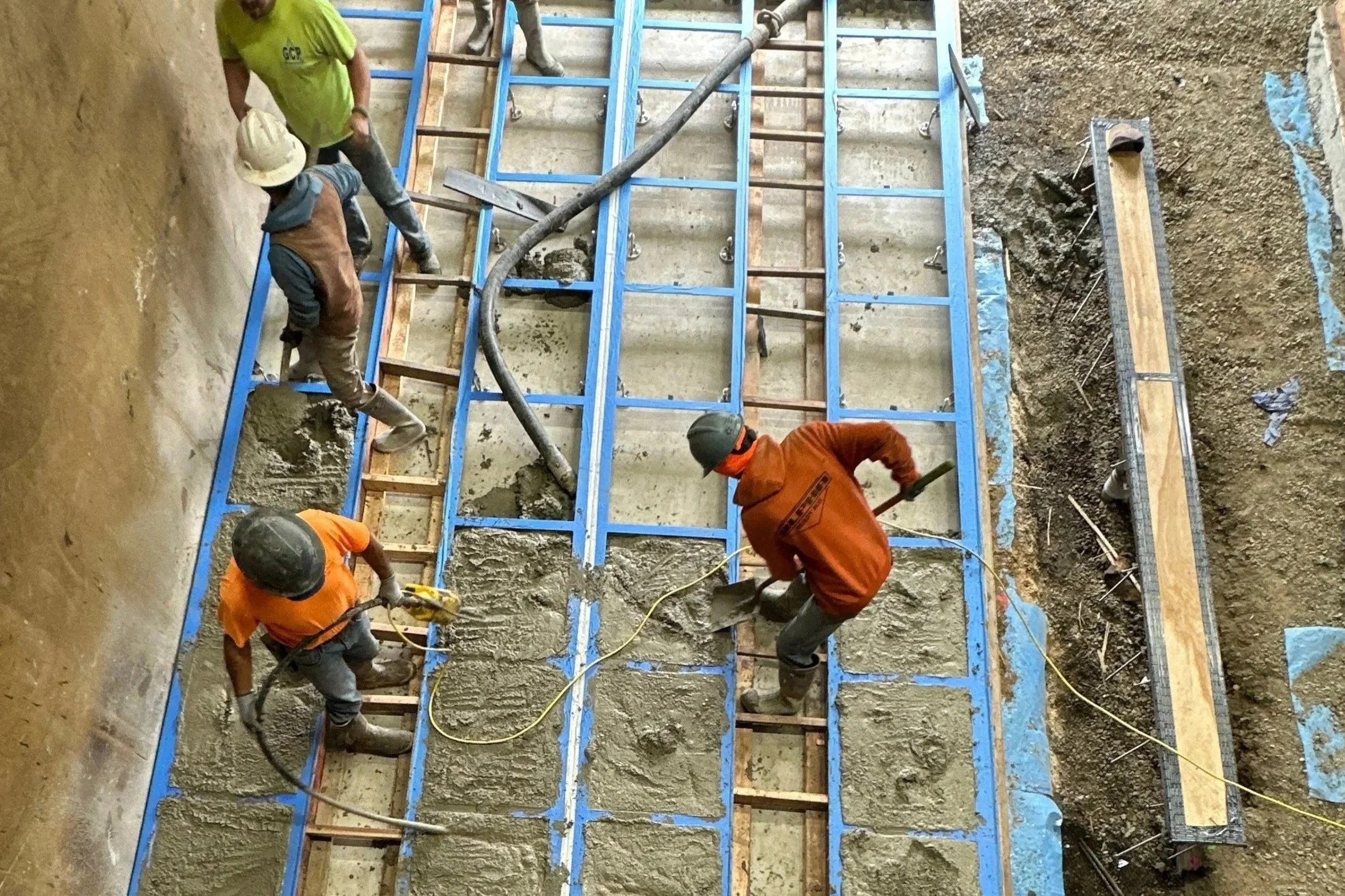 Workers pouring concrete into a construction form for a building foundation, using a concrete hose and shovels, with some workers wearing safety helmets.