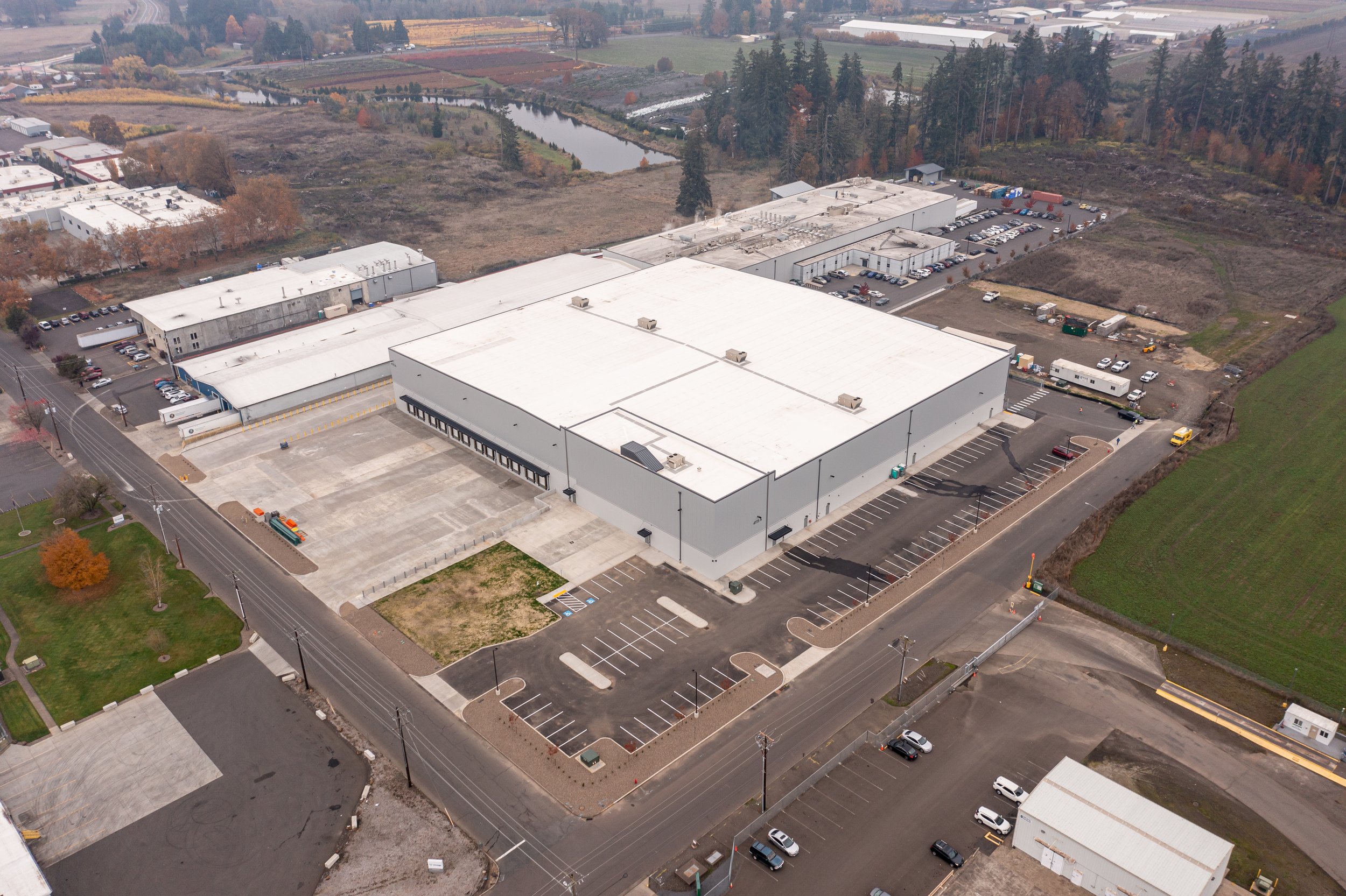Aerial view of a large commercial warehouse building with a parking lot, surrounded by roads and open land with some trees and fields.