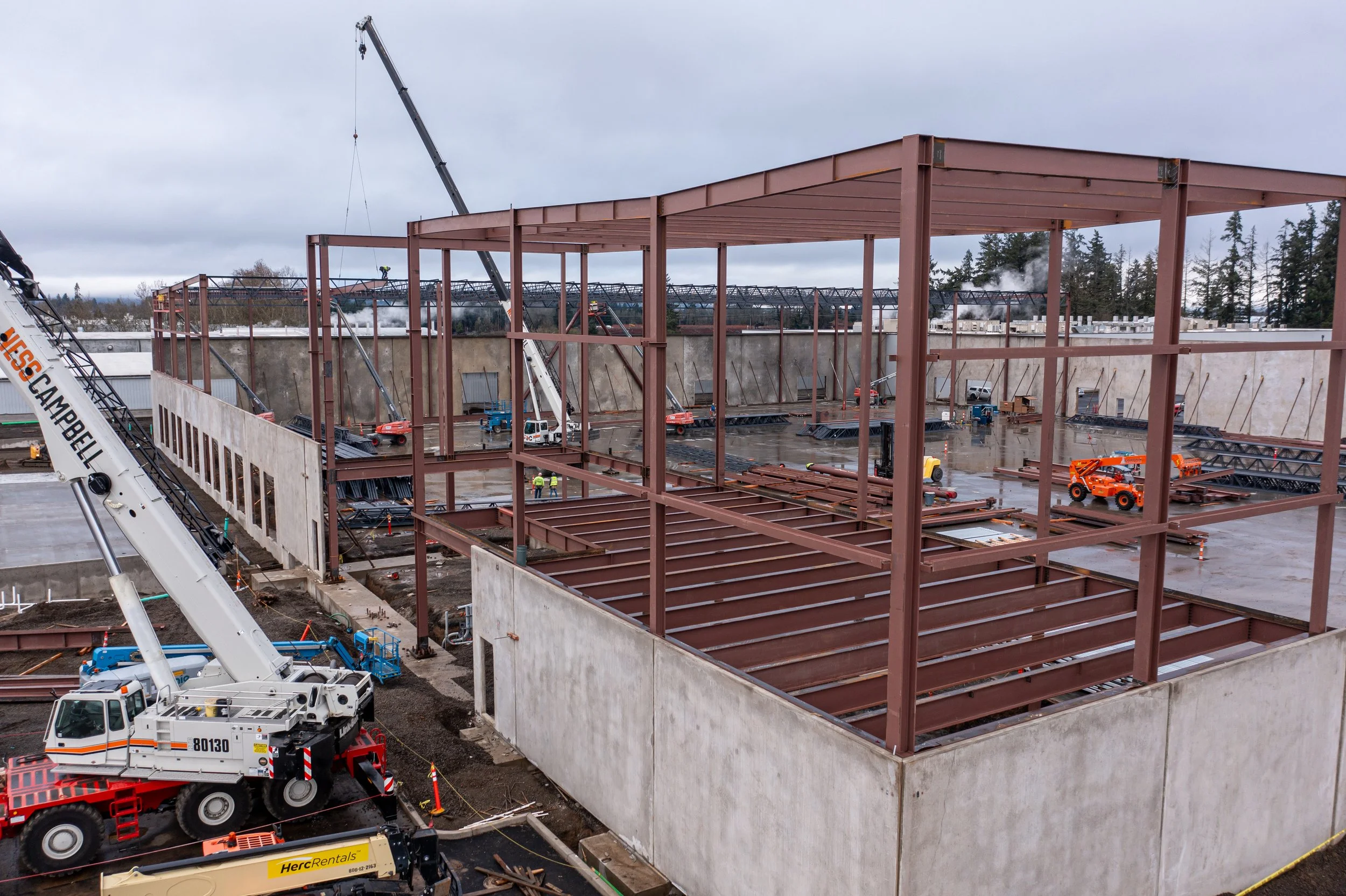 Construction site with a steel framework for a building, cranes, and construction vehicles on a cloudy day.