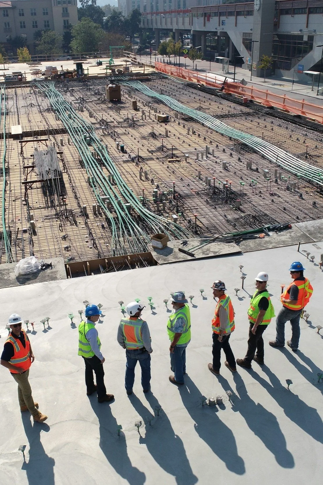 Construction workers wearing safety vests and helmets standing on a concrete floor observing a construction site with rebar and electrical wiring above.