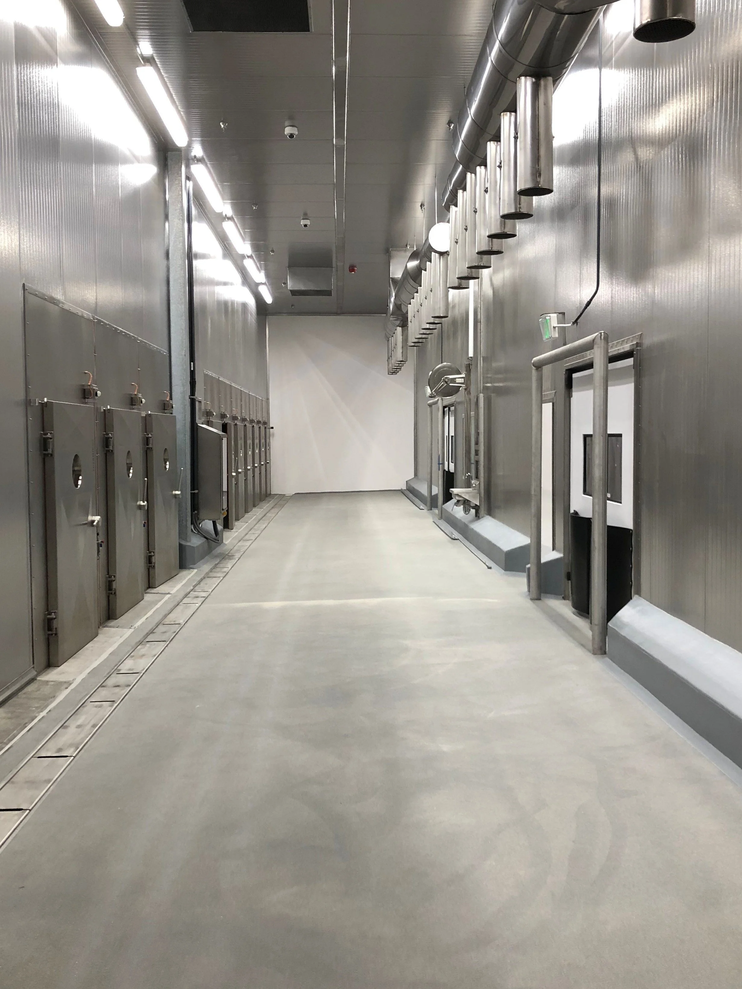 Empty industrial corridor with stainless steel lockers on left, large ventilation pipes on right, and a gray concrete floor.