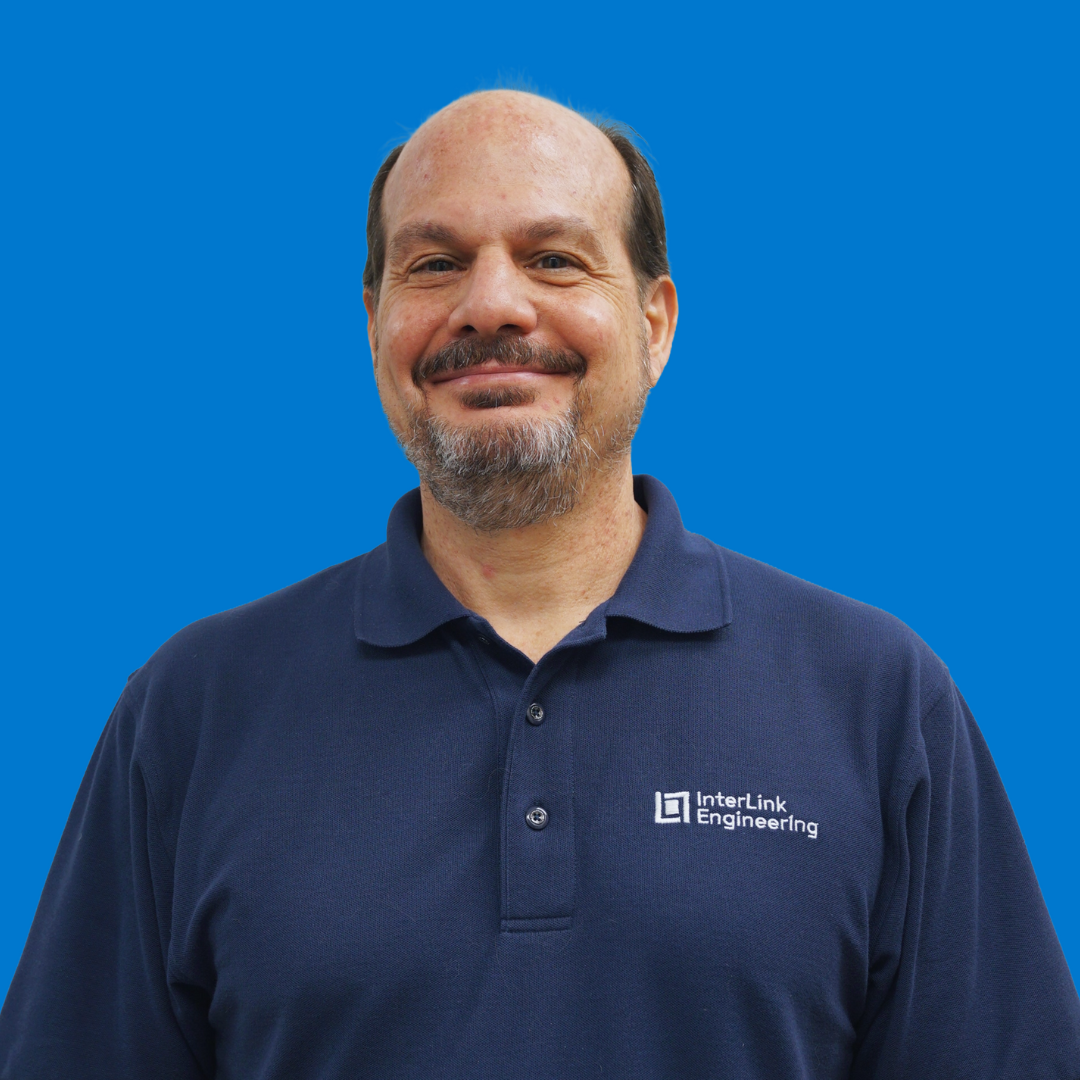 A man with a beard smiling, wearing a navy polo shirt with the InterLink Engineering logo, against a solid blue background.