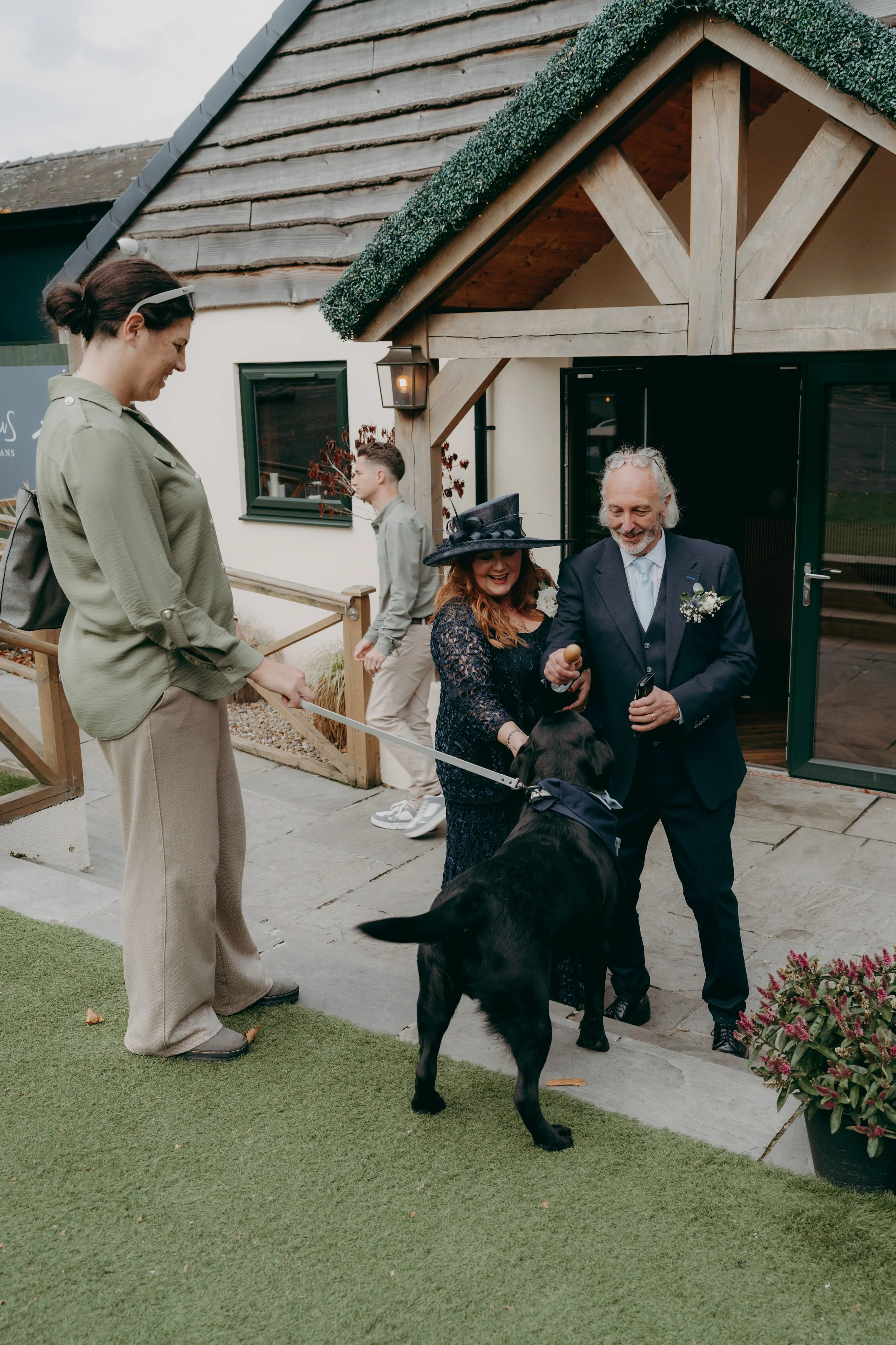 People greeting a black Labrador dog outside a building at a gathering or event, with a woman in a black dress and hat, an older man in a suit, and a woman holding the leash.