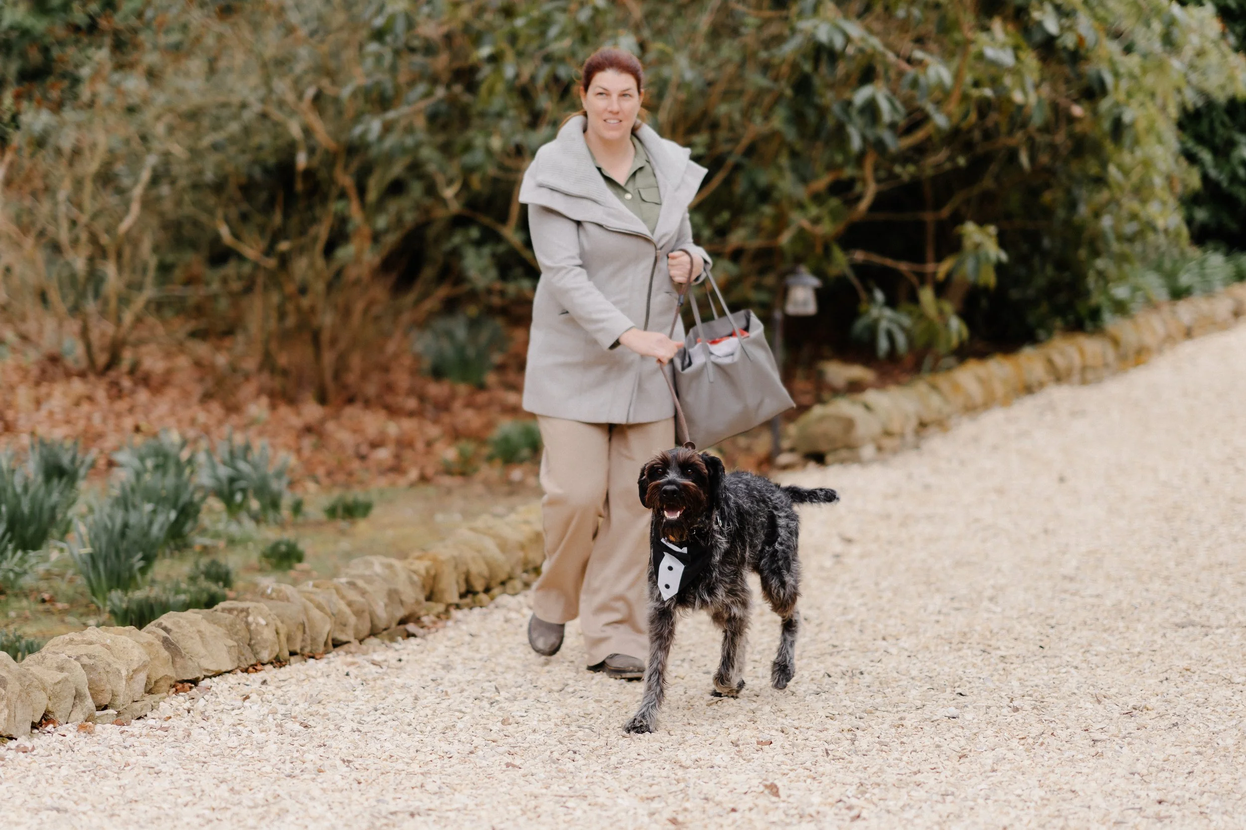 A woman walking her dog on a gravel path surrounded by bushes and plants.