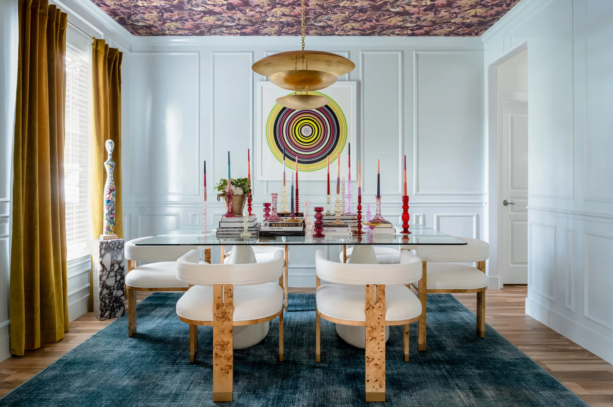 A modern dining room with a white table, six white chairs, a blue area rug, and a collection of colorful candlesticks and books as a centerpiece. There is abstract artwork on the wall, a tall decorative vase near the window, and gold curtains on the left side. The ceiling has a floral pattern.