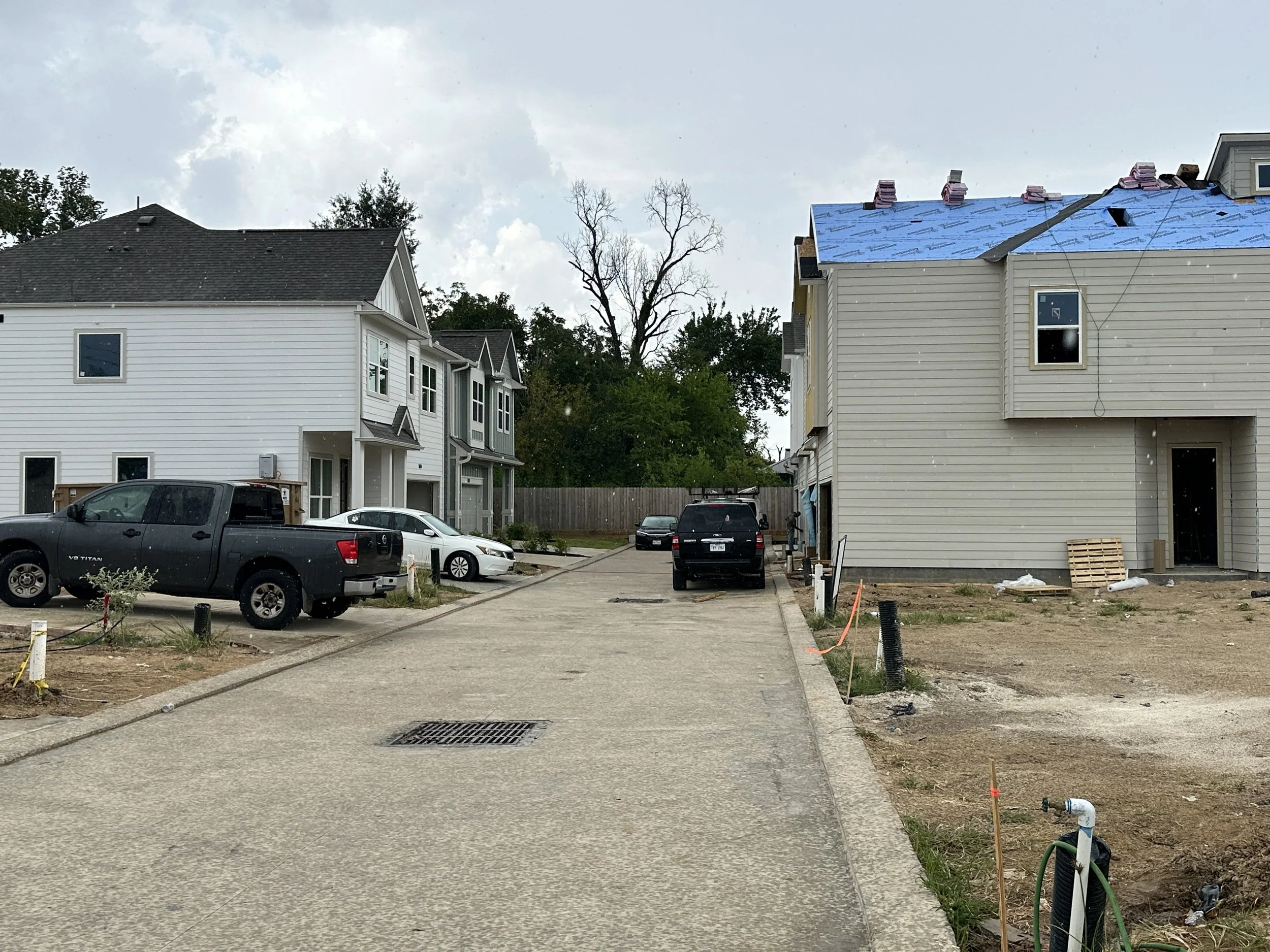 Two houses are under construction, with one house having a blue roof and construction materials on top. The area around the houses has cars parked and construction equipment, with visible construction work such as pipes and materials on the ground.