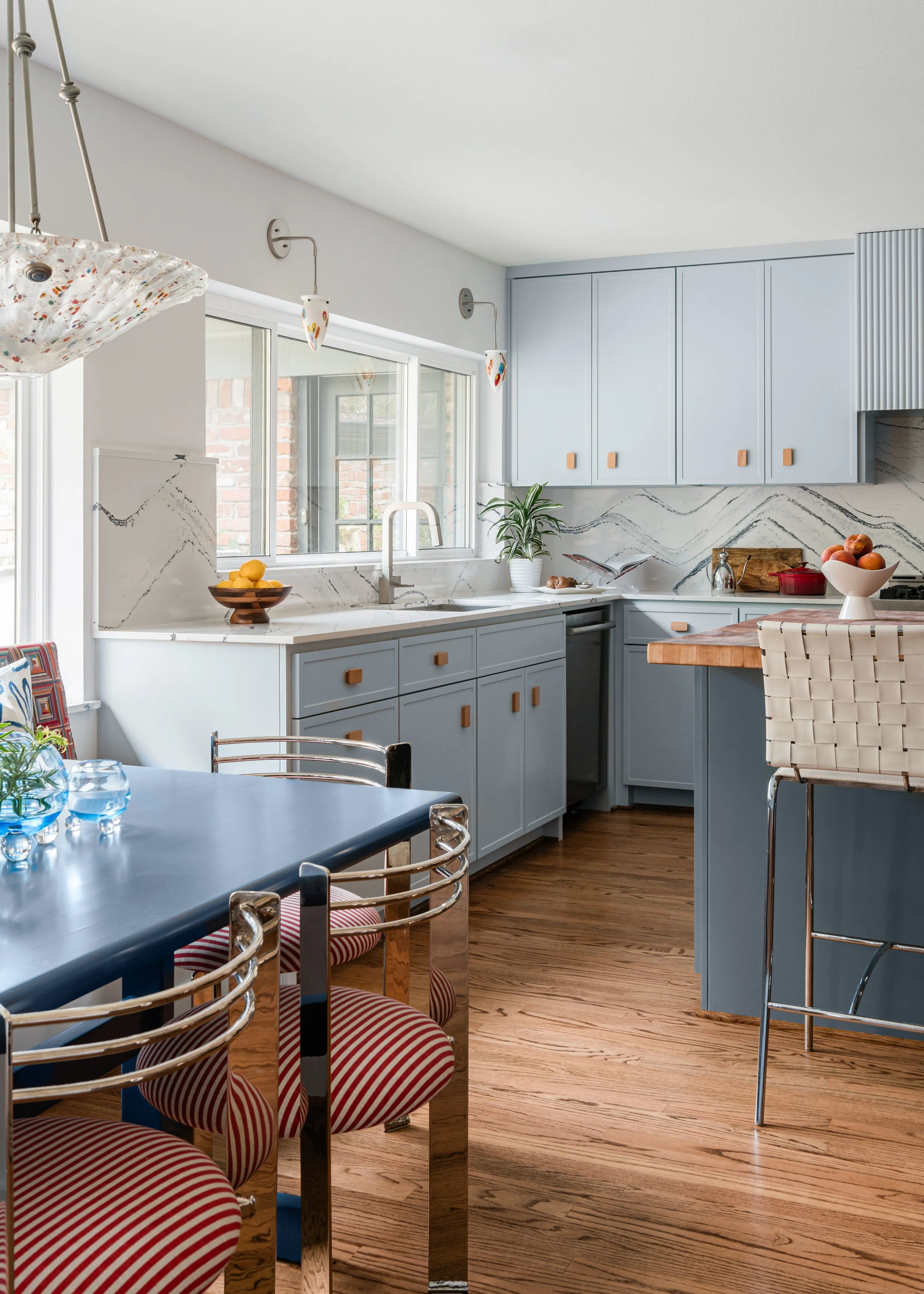 Modern kitchen with blue cabinets, wooden accents, marble backsplash, and a dining table with striped red and white cushioned chairs.