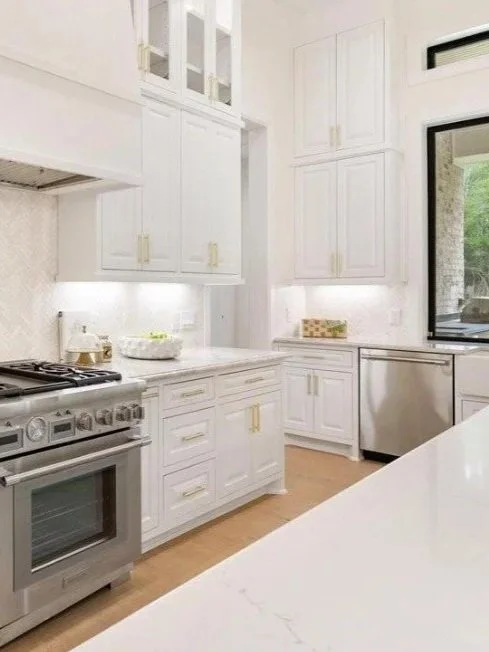 Modern white kitchen with white cabinets, a stainless steel oven, a white countertop, a large window, and under-cabinet lighting.