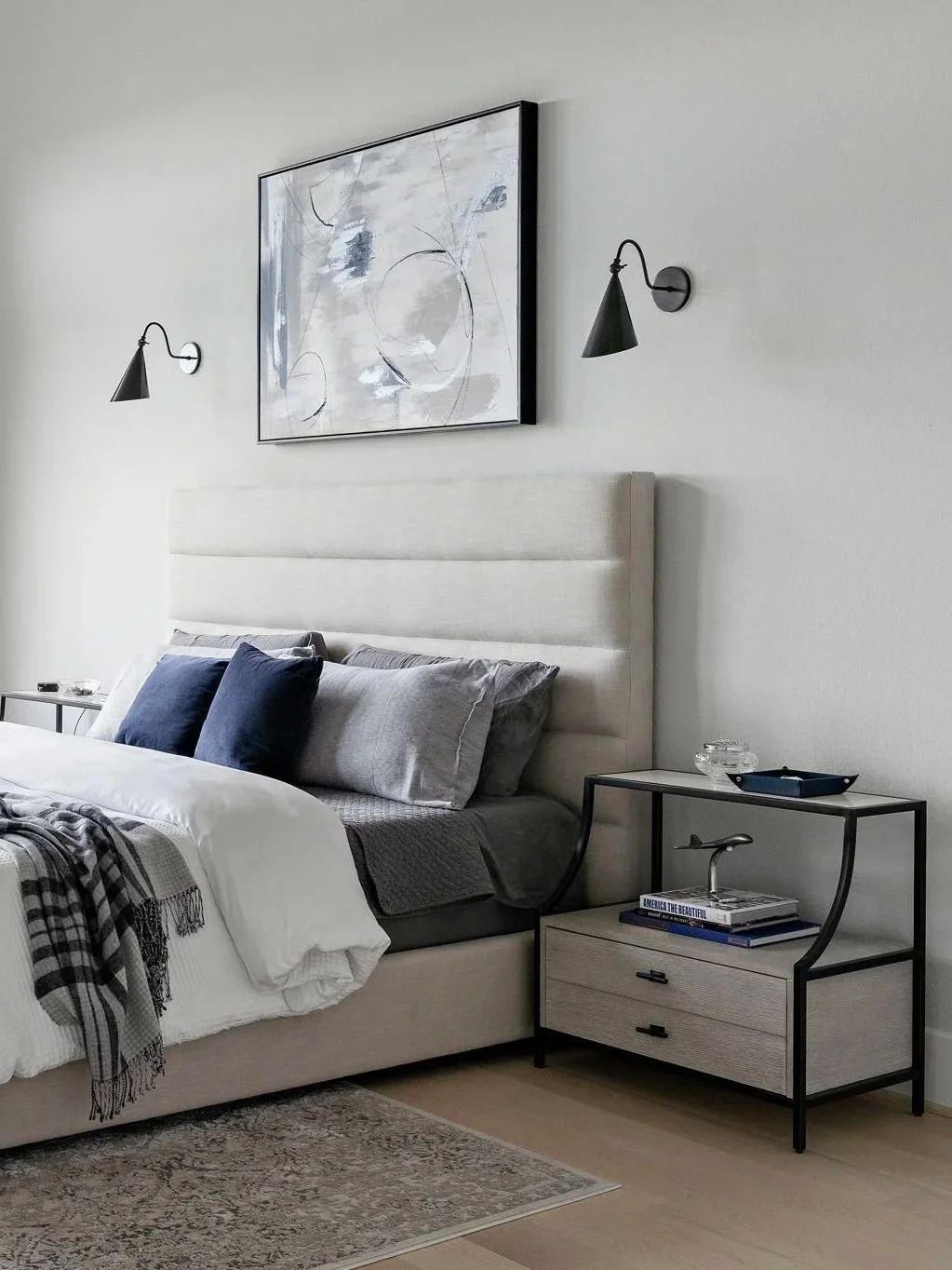 Modern bedroom with beige upholstered bed, gray and navy bedding, abstract black-and-white artwork, wall-mounted black lamps, and a small nightstand with books and decorative glass items.