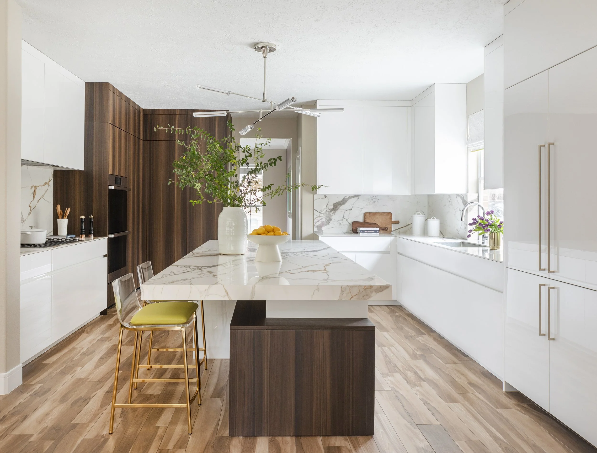 A bright kitchen with white and wood cabinets, marble countertops, and a large island. Then, a vase of green plants and a bowl of oranges add fresh color.