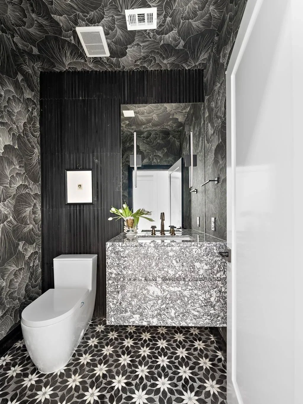 Modern bathroom with black floral wallpaper, black wood paneling, and patterned floor tiles; features a white sleek toilet, a grey and white marble vanity with a vase of flowers, and a large mirror.