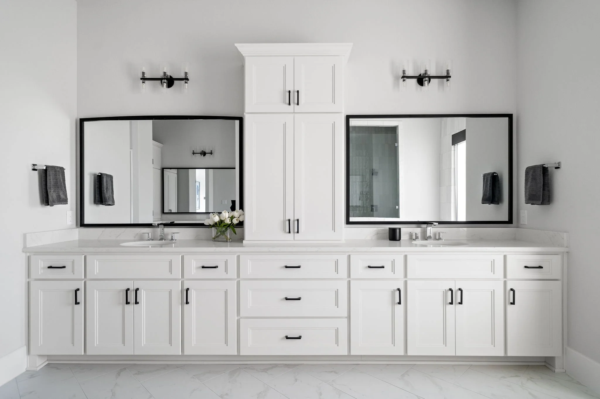 Modern white bathroom vanity with two sinks, large mirrors, black hardware, black wall sconces, and a white cabinet in the center.