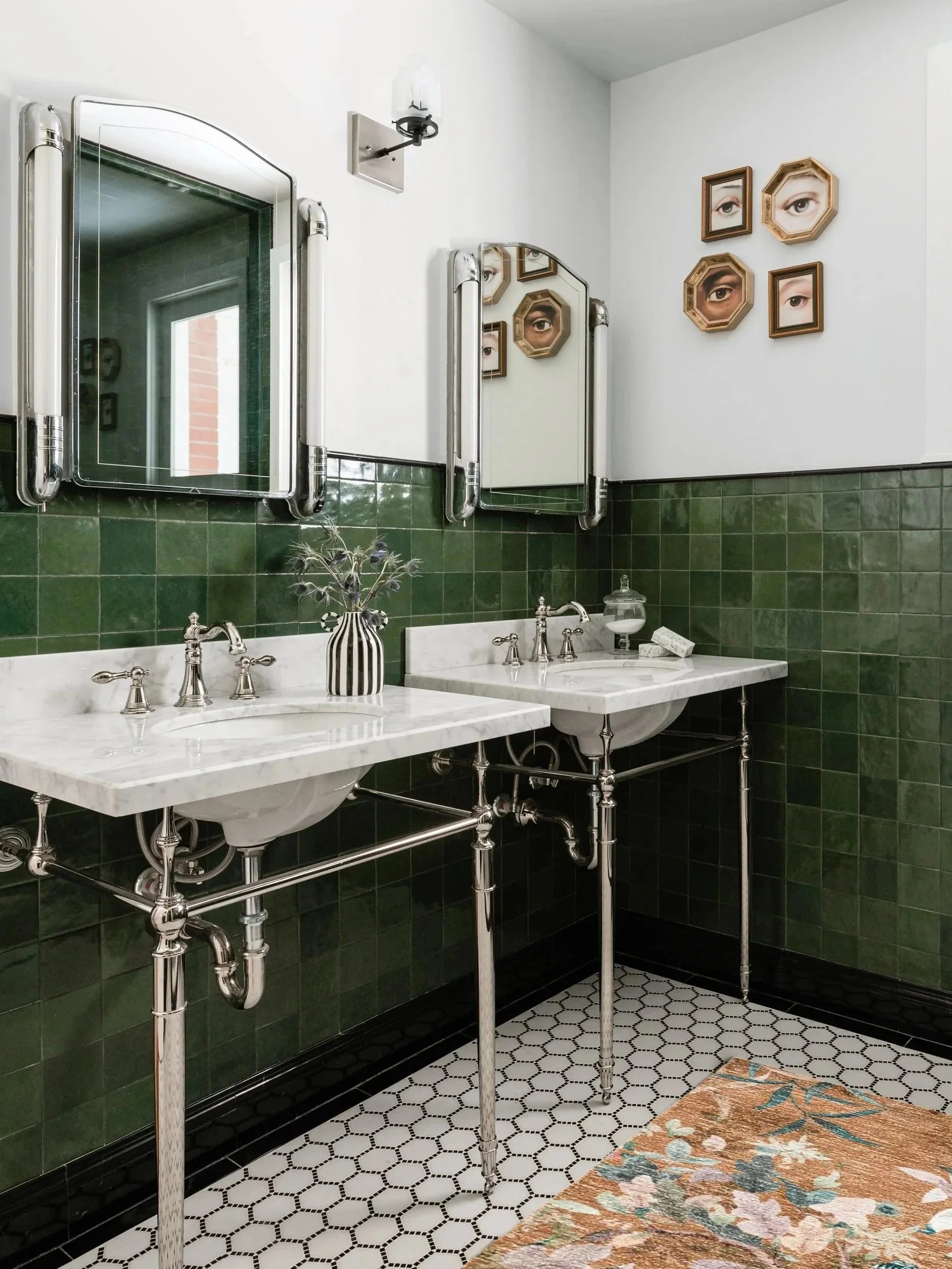 Vintage bathroom with dual white marble sinks, green tiled walls, hexagon black-and-white tiled floor, and framed eye artwork on the wall.