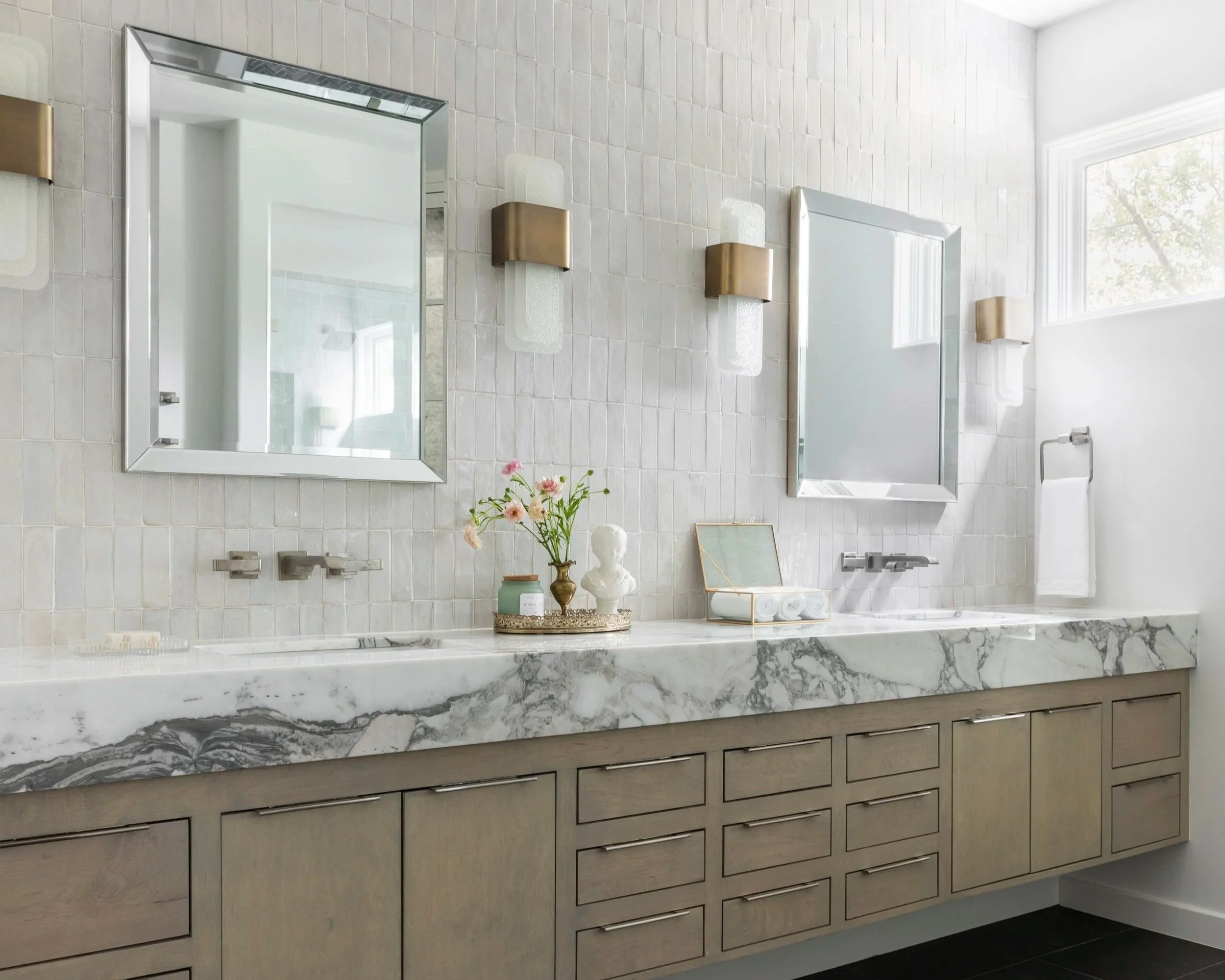Modern bathroom vanity with a marble countertop, two large mirrors, vase of flowers, decorative bust, and wall-mounted lights.