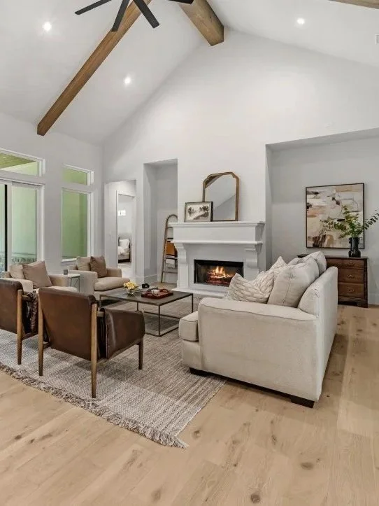Living room with high vaulted ceiling, white walls, wood beams, large windows, white and beige furniture, fireplace, and wooden flooring.