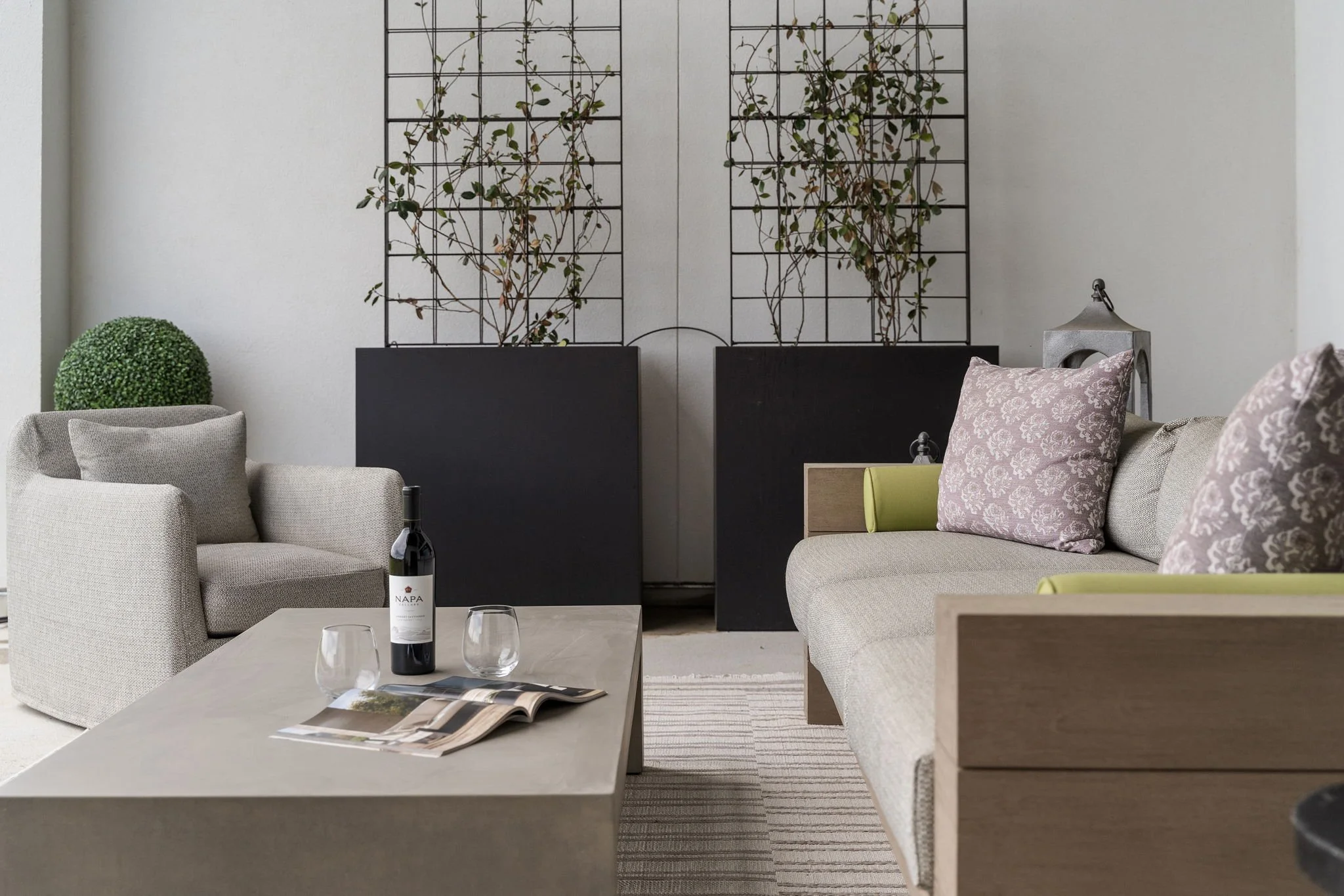 Interior of a living room with light-colored upholstered sofas, decorative pillows, a coffee table with a wine bottle, glasses, and magazine, a white wall with black metal wall art featuring vines, and a small green shrub on a side table.