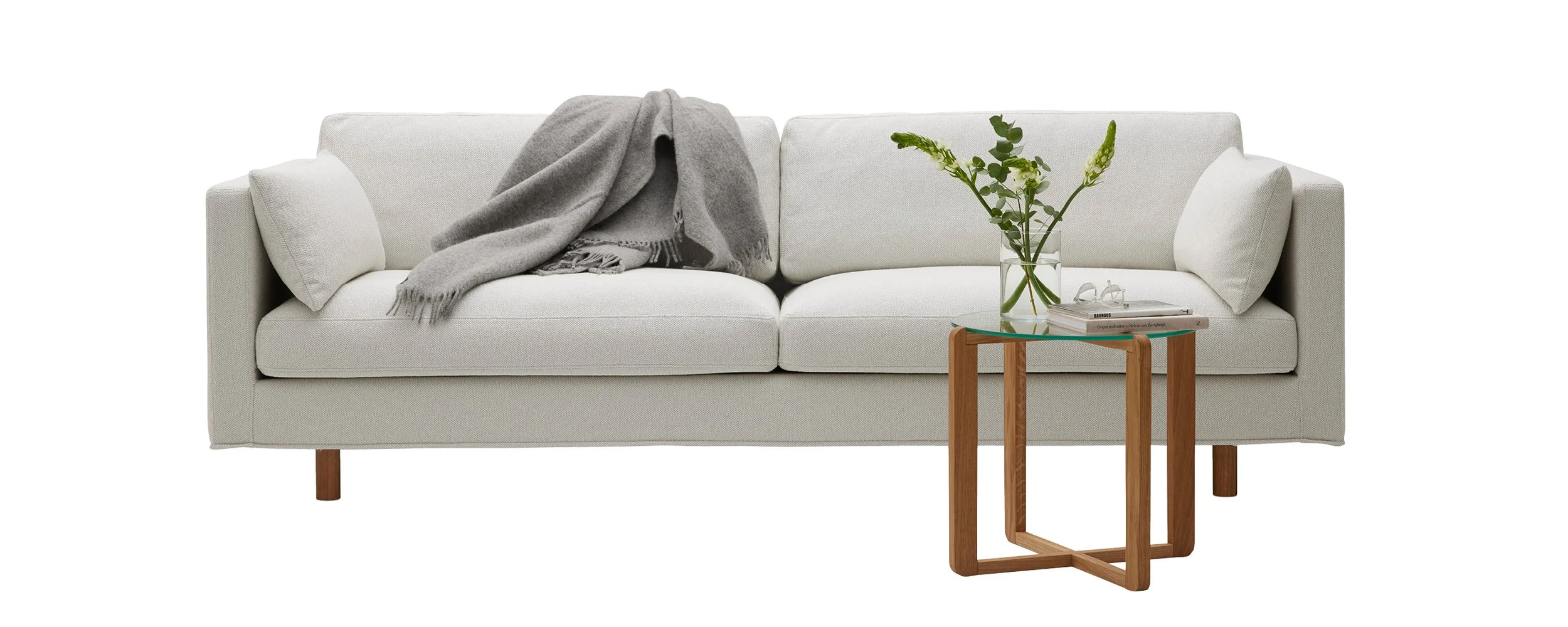 Vit soffa med grå filt över ryggstödet och ett sidobord i trä med glasskiva, dekorerat med en vas med gröna blommor och en bok.