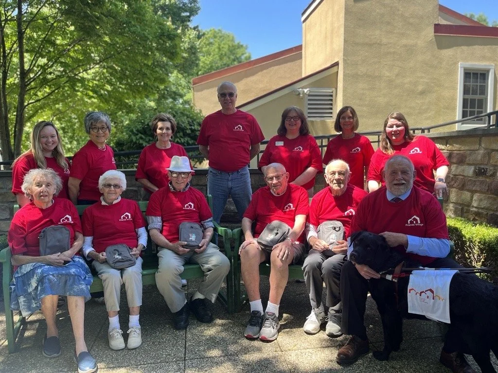 Group of people, mostly elderly and middle-aged women, and a few men, sitting and standing outside on a sunny day, all wearing red shirts with a white house logo, some holding small bags, and one person with a service dog.